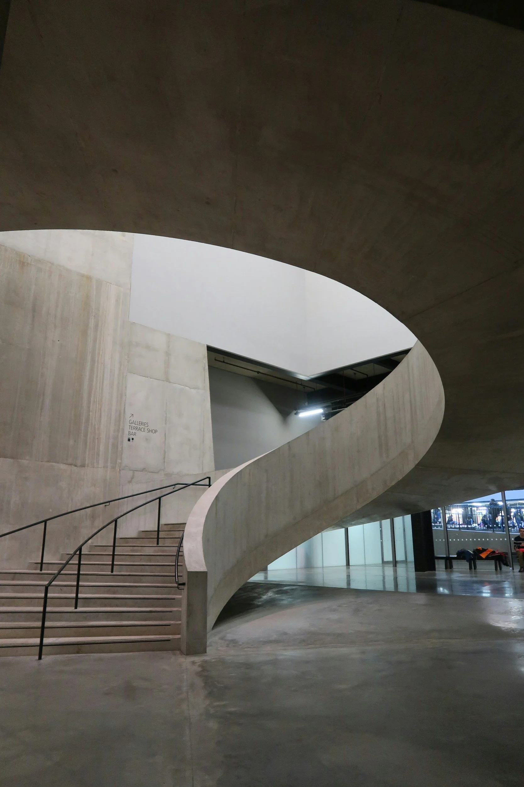 Interior view of a modern building with a curved staircase, concrete walls, and an open ceiling with visible insulation.
