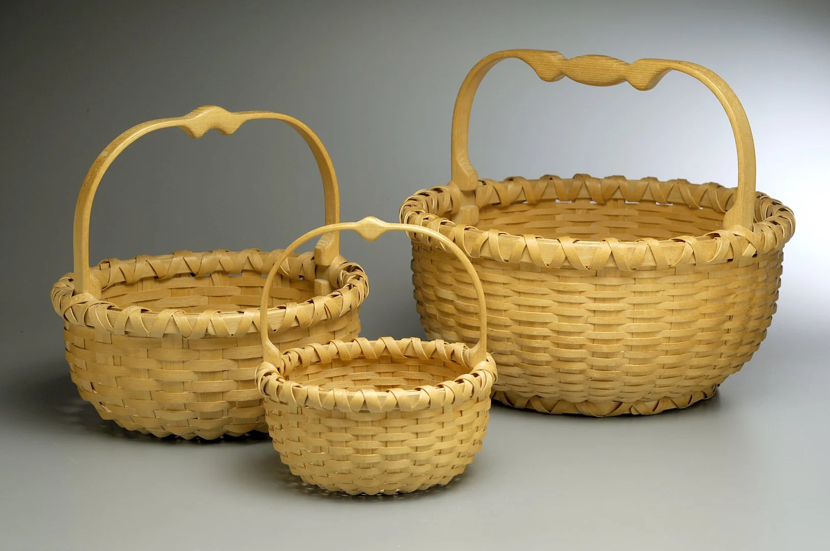 Three woven wicker baskets of varying sizes with curved handles, placed on a plain surface with a gradient background.