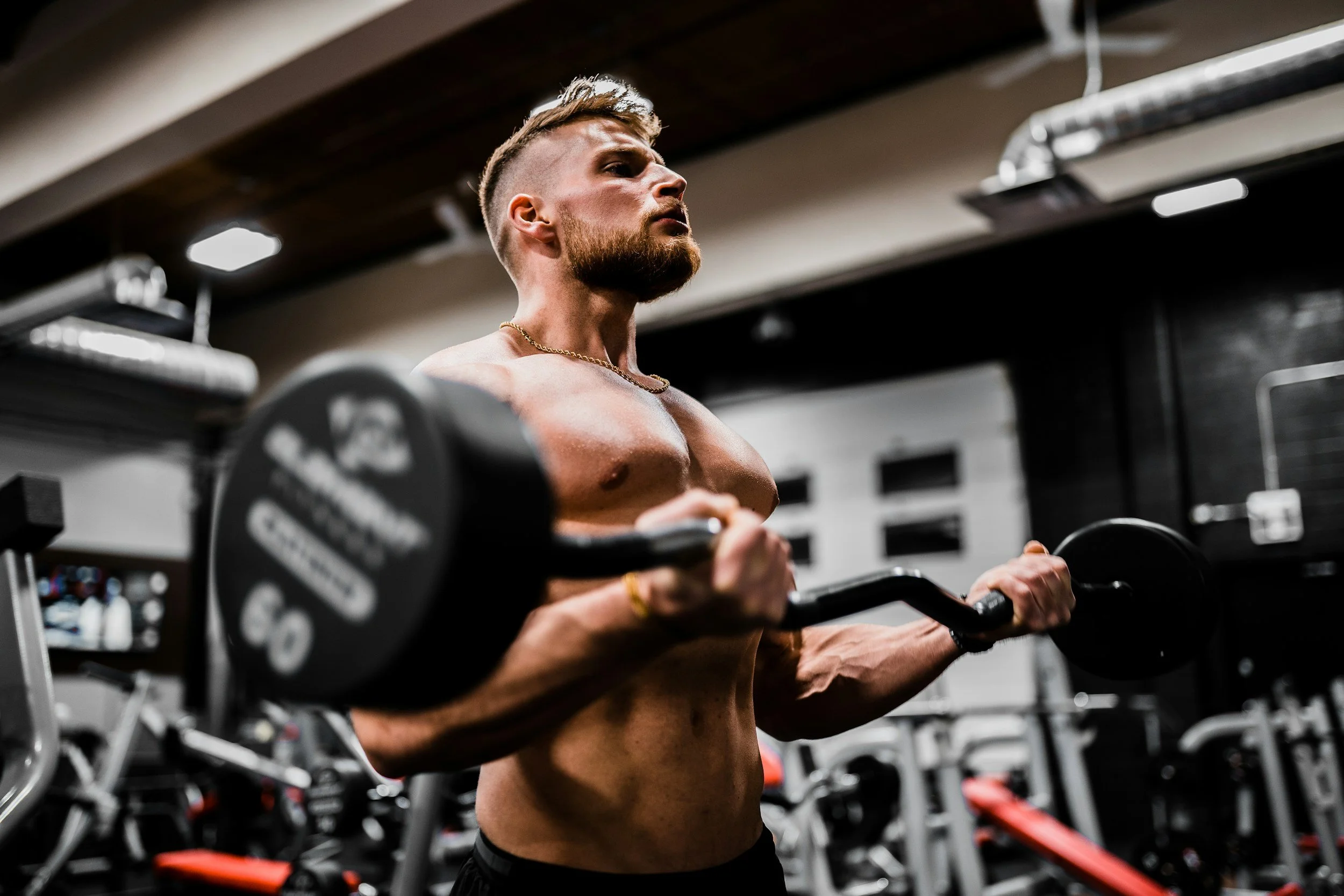 A shirtless man with a beard and short hairstyle is lifting a dumbbell at the gym