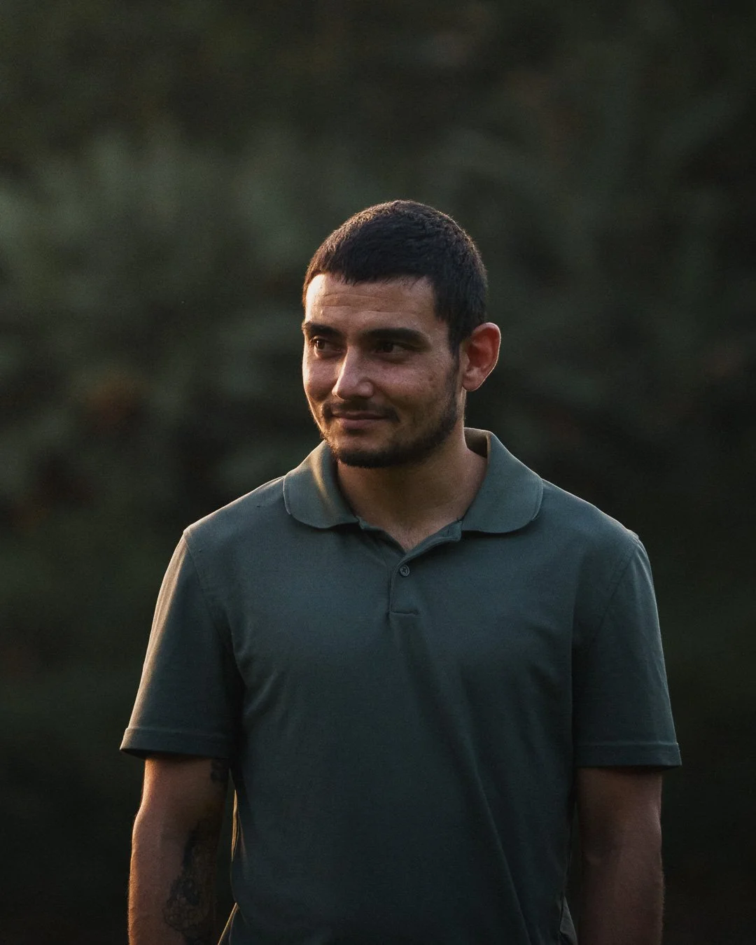 A young man with short dark hair, a beard, and a tattoo on his left arm, wearing a dark green polo shirt, standing outdoors during sunset with a blurred natural background.