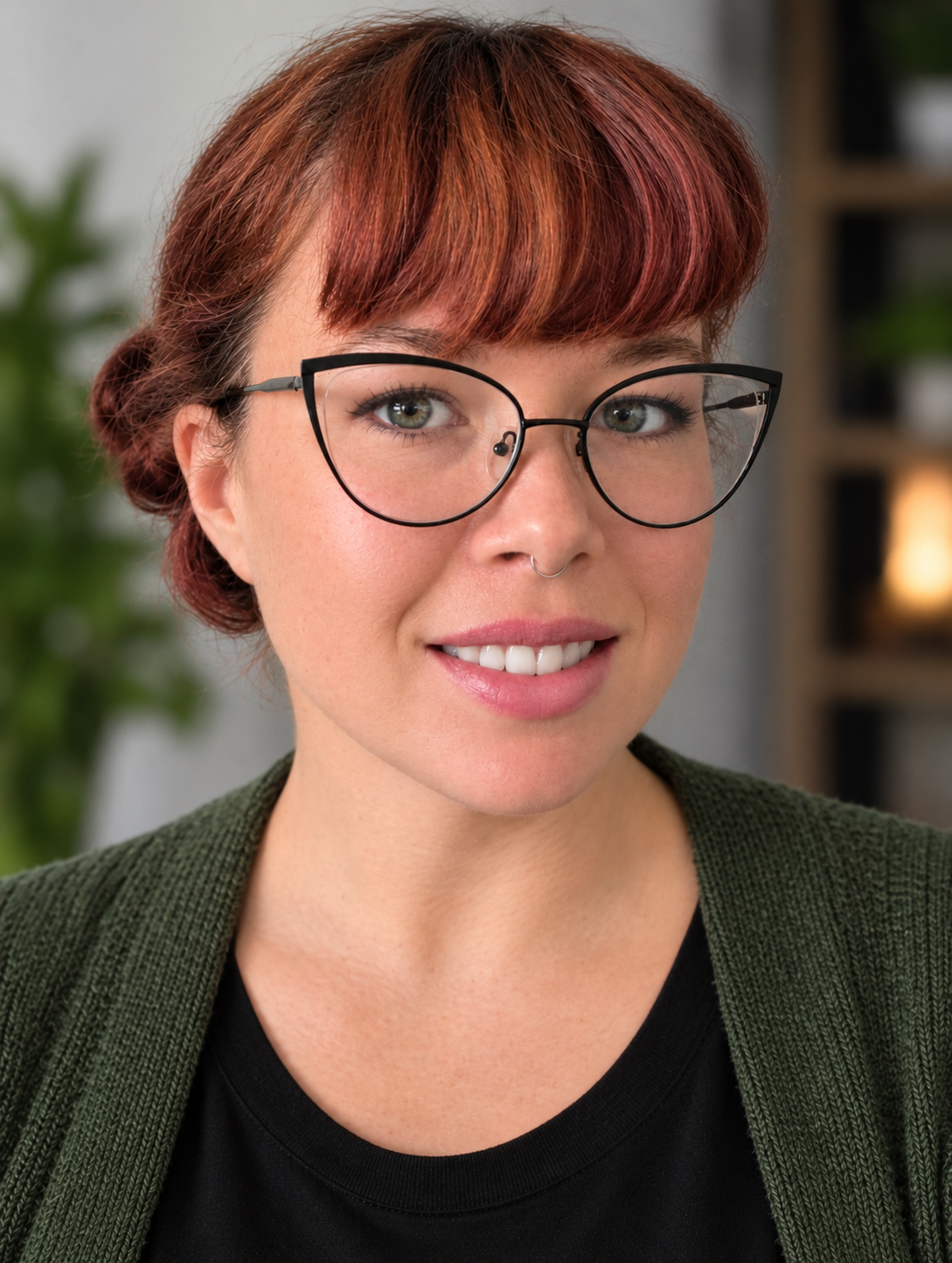 Close-up portrait of a woman with short red hair, wearing glasses, a septum piercing, and a black top with a green cardigan, smiling indoors.
