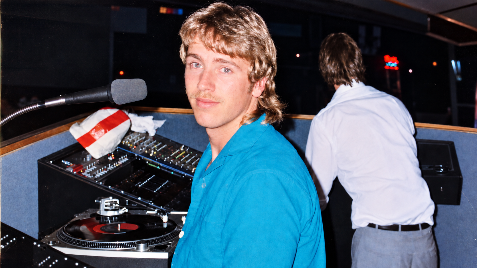 A young man with blonde hair and a mustache in a blue shirt sitting at a DJ booth, looking at the camera, with another man in a white shirt facing away in the background.