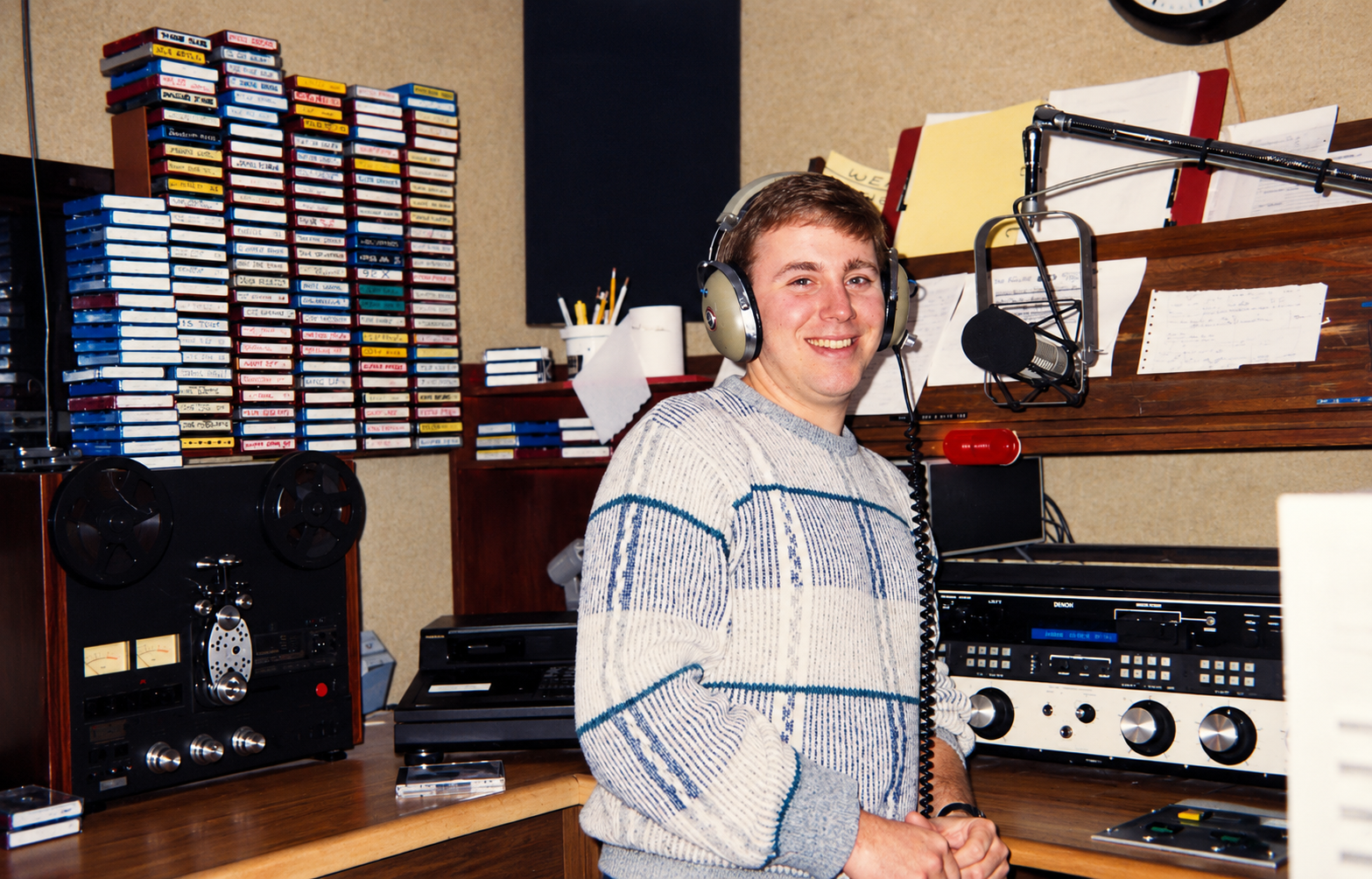 A young man in a radio station studio wearing headphones and smiling at the camera, with audio equipment and cards with notes on the wall behind him.