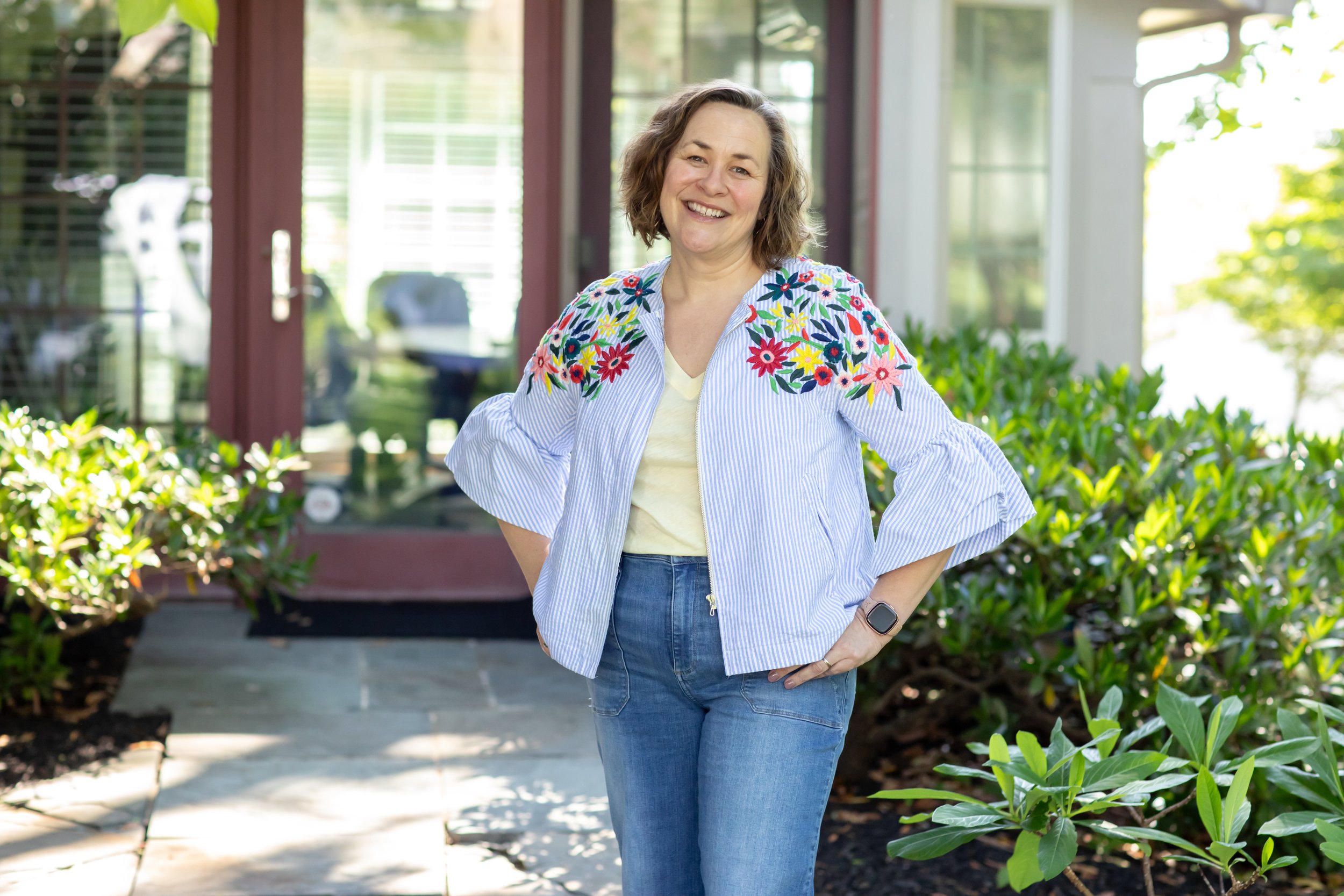 Smiling woman in casual attire standing outdoors in a garden area, with plants and a porch in the background.