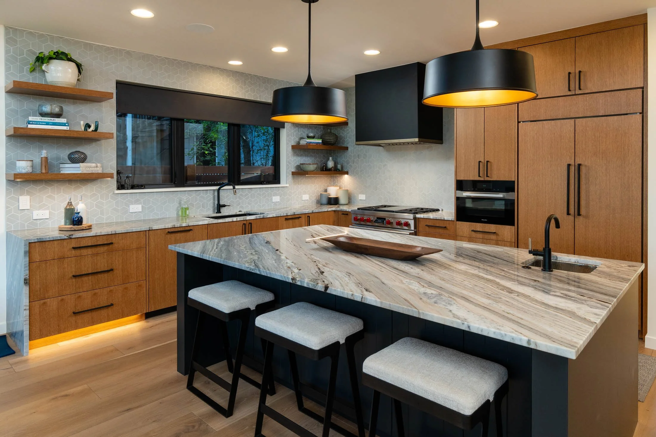 Modern kitchen with wooden cabinets, a marble countertop island, black pendant lights, and floating shelves with decor, featuring a window with a view of trees outside.