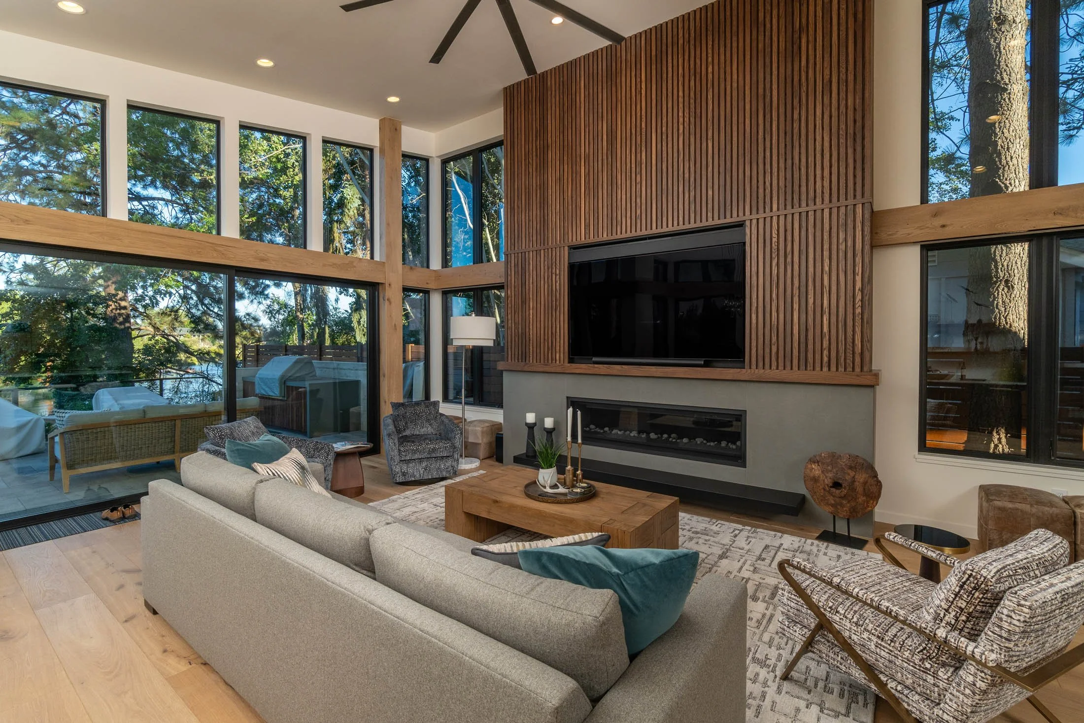 Interior view of a modern living room with large windows showing trees outside, a fireplace with wood paneling, a ceiling fan, and various seating options including a sofa and chairs.