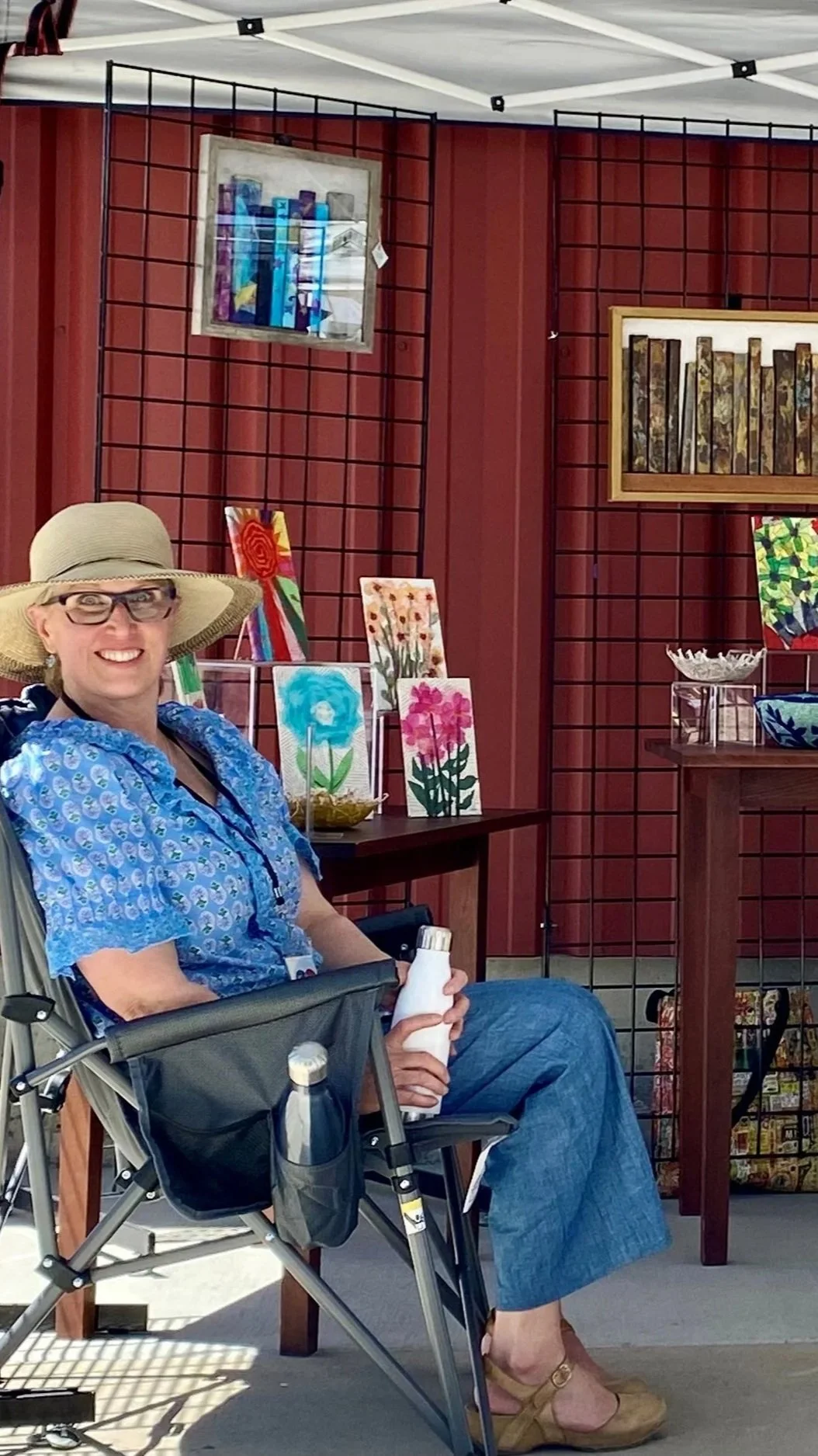 Adriane Herrick Juarez library director and artist in Park City, UT wearing glasses and a wide-brimmed hat sitting in a chair, smiling, at an art display booth with colorful paintings and decorative upcycled book art on display.