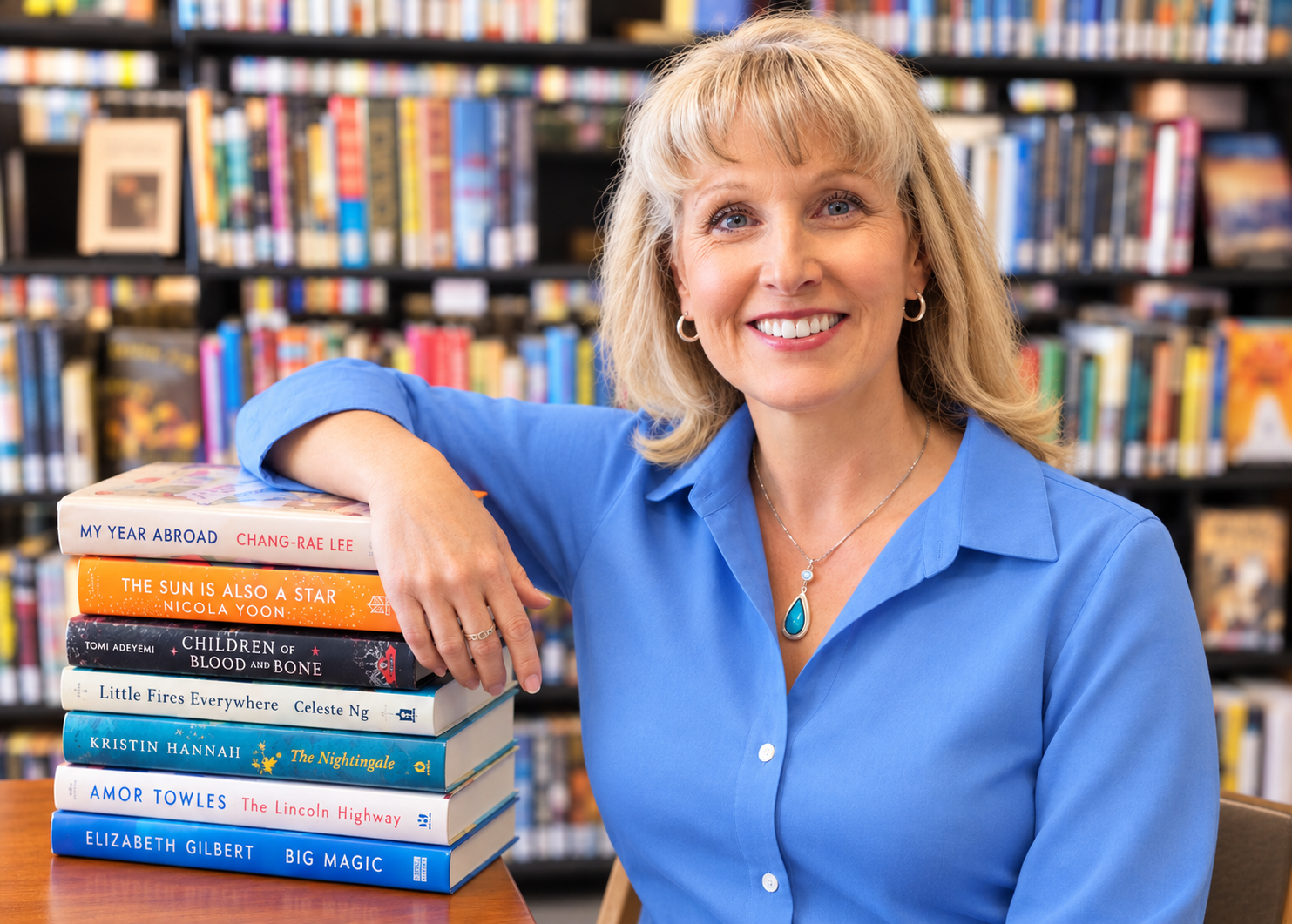 A woman with blonde hair and blue eyes sitting in a library, wearing a blue shirt, smiling, with a stack of books on the table beside her.