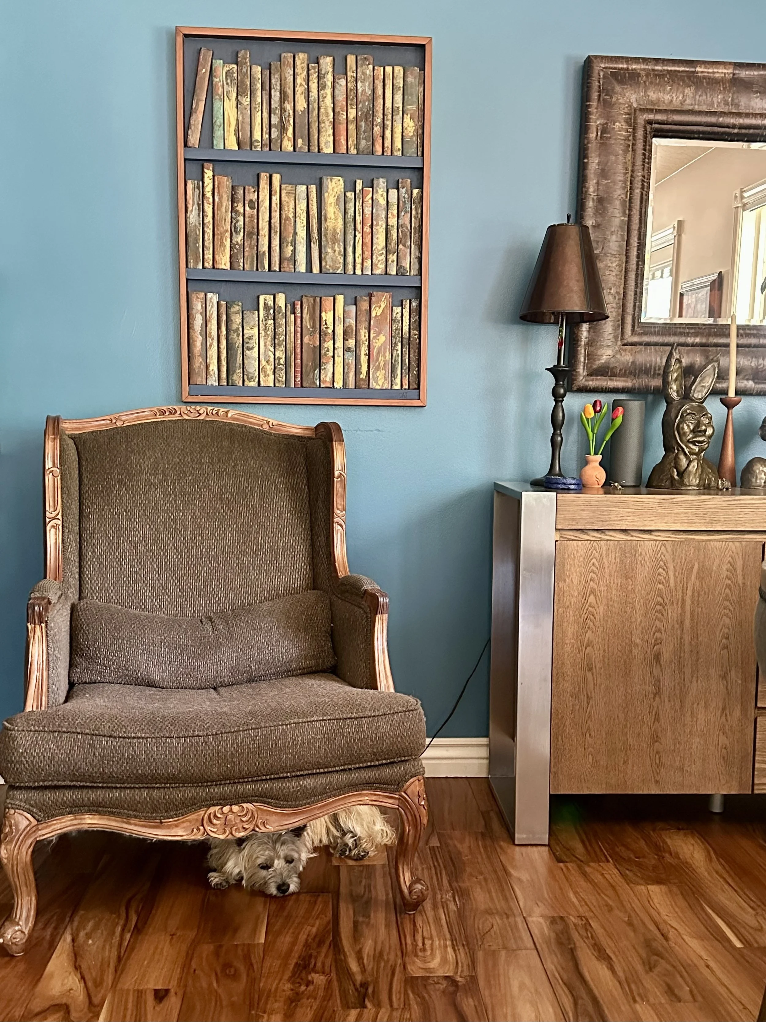A cozy room with a vintage armchair, a dog lying underneath, a blue wall, a bookshelf with vintage books, a large ornate mirror, a black table lamp, a small vase with tulips, a decorative sculpture, and a tall candlestick.