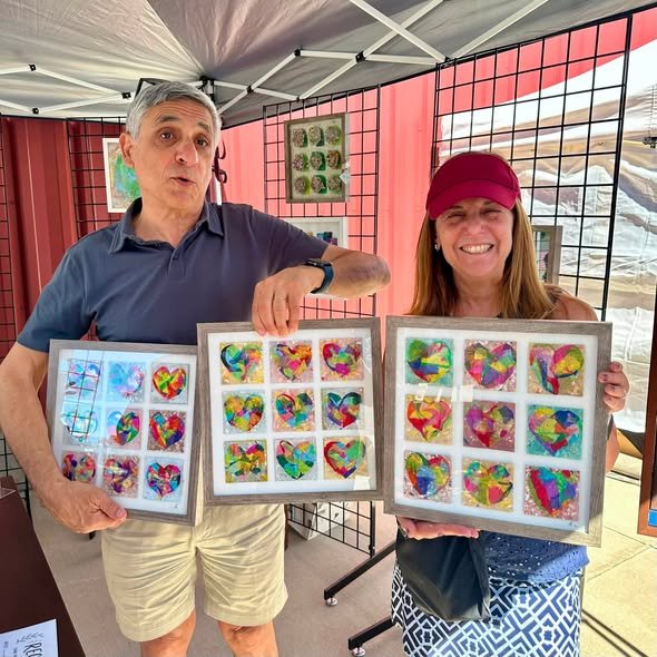 A man and woman standing indoors under a canopy, each holding frames with colorful heart-shaped crafts made from small, multicolored pieces. The man wears a blue polo shirt and beige shorts, and the woman wears a red cap and patterned pants, both smiling.