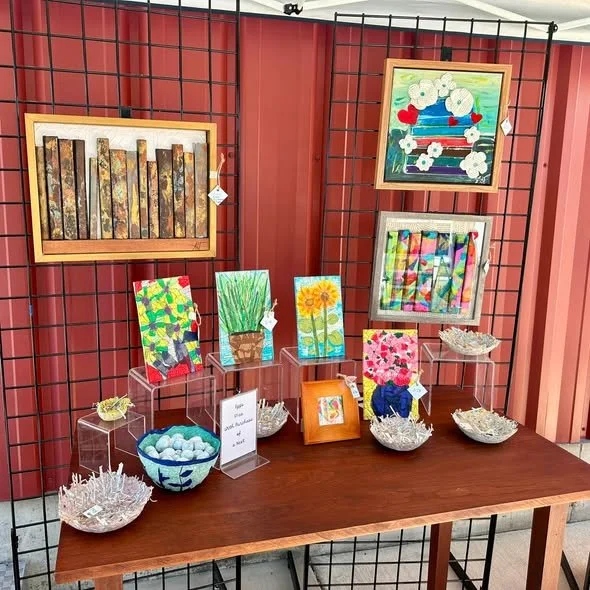 Display of framed paintings and small colorful artwork on a table and grid backdrop, with bowls of decorative items and small signs.