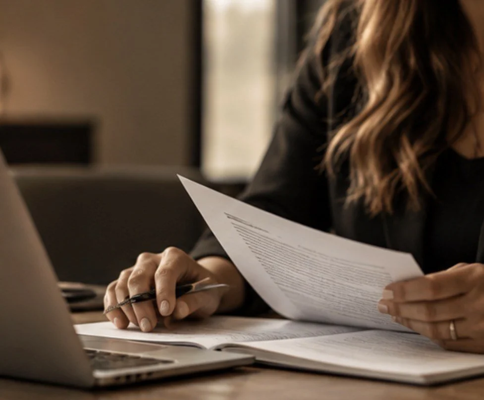 A woman sitting at a desk, holding a sheet of paper, with a laptop and notebooks in front of her.