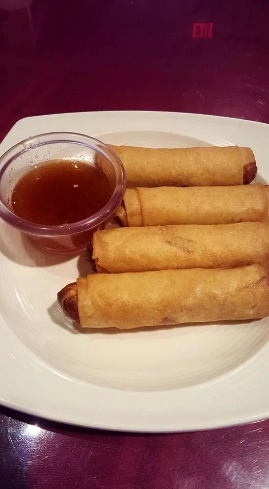 Four fried spring rolls with a cup of dipping sauce on a white plate.