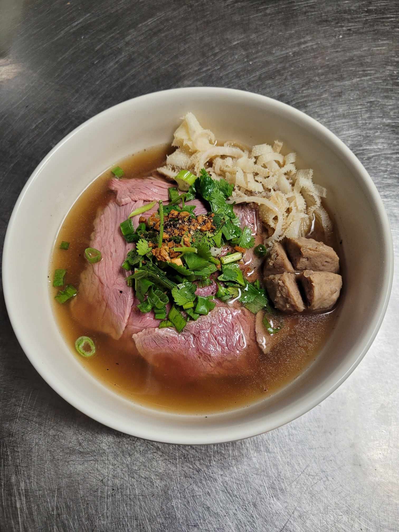 A bowl of Vietnamese pho with slices of beef, noodles, green onions, cilantro, and broth, on a stainless steel surface.