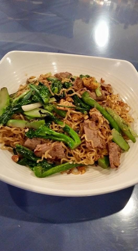 A bowl of stir-fried noodles with beef and green vegetables on a dark table.