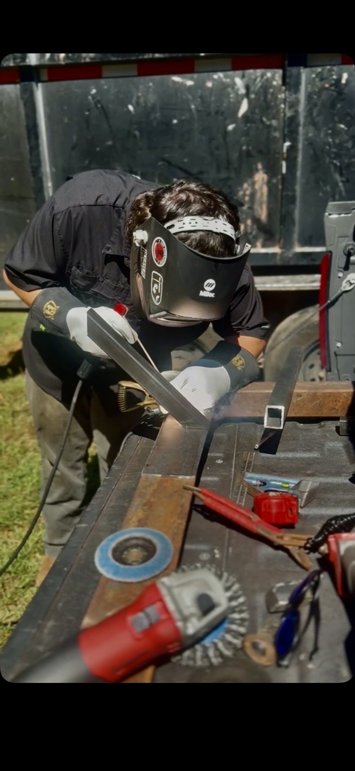 Person welding a metal piece outdoors with safety gear, surrounded by tools and equipment.