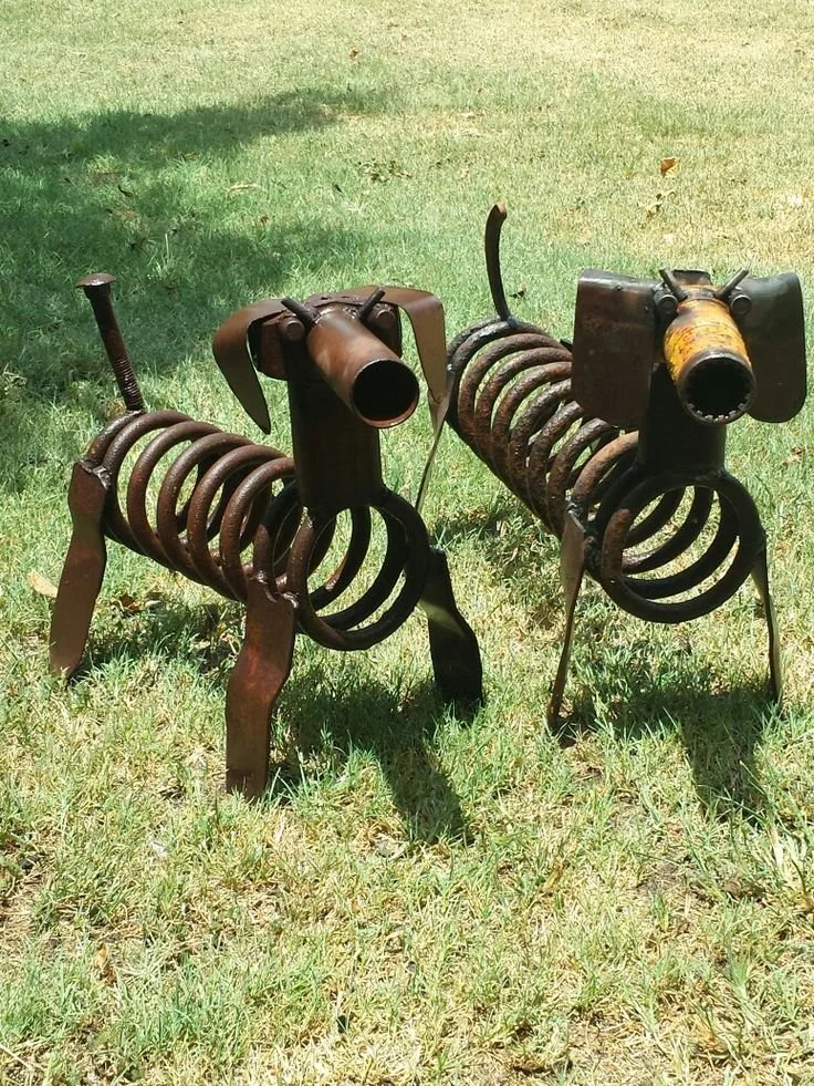 Two metal sculptures resembling dogs made from springs, pipes, and scrap metal, standing on a grassy area.