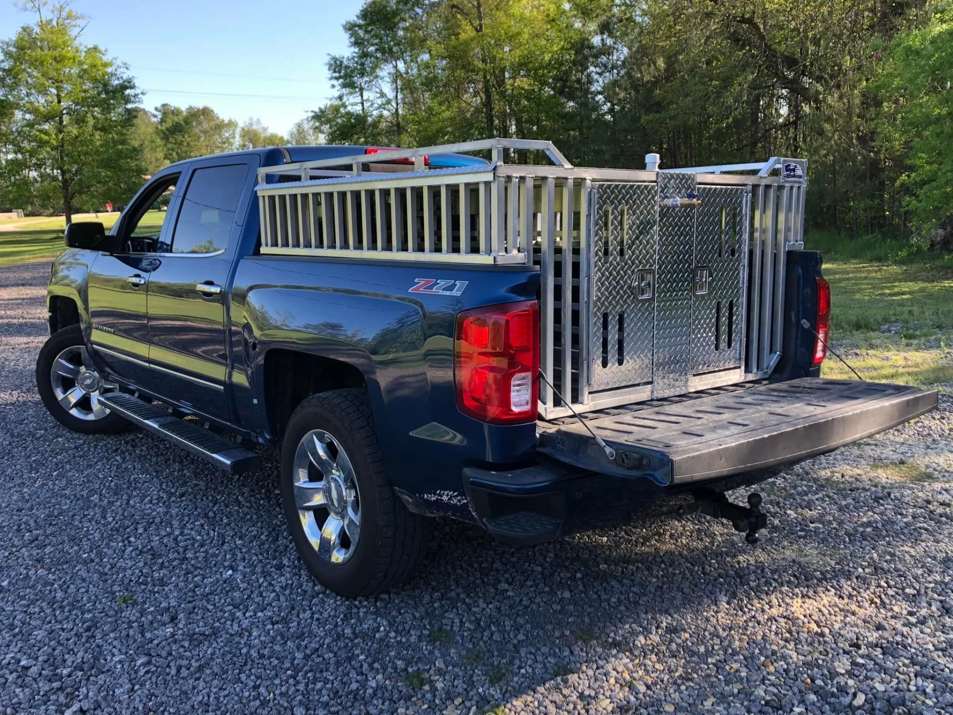 Black pickup truck with dog kennels in the truck bed, parked on a gravel surface, with trees and blue sky in the background.