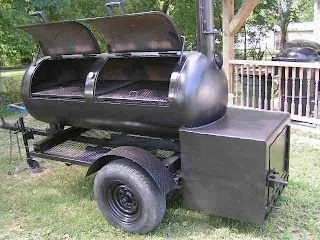 A large, barrel-shaped smoker grill mounted on wheels with a side firebox, outdoors on grassy area with trees and a wooden fence in the background.
