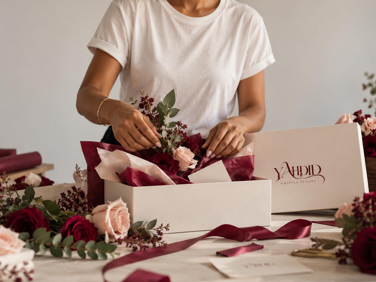 Florist arranging red and blush roses in a gift box with burgundy ribbon and floral wrapping supplies