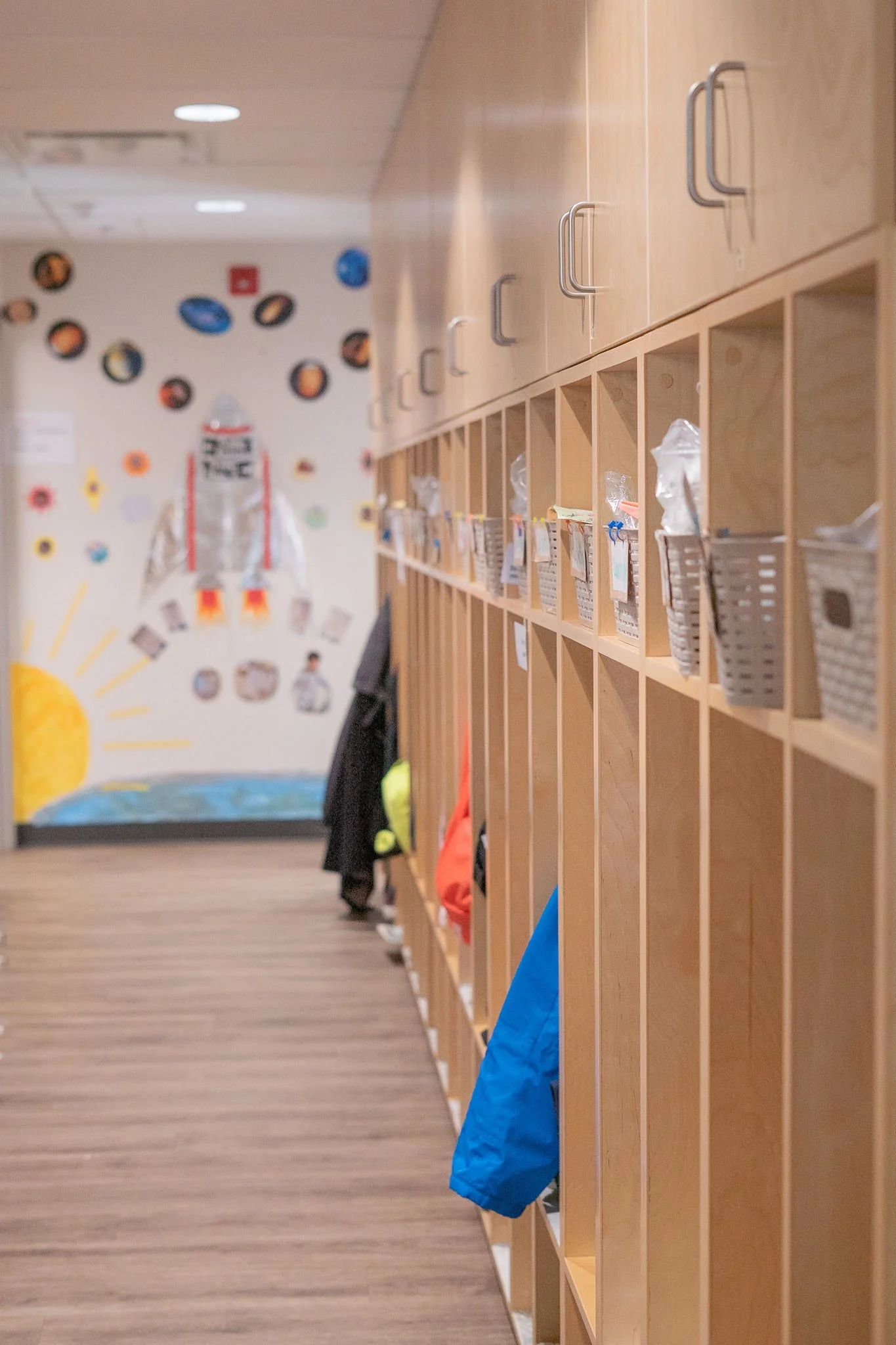 A hallway in a daycare with wooden cubbies and hooks on the right side, some jackets hanging. The background shows a wall decorated with colorful children's artwork and cutouts.