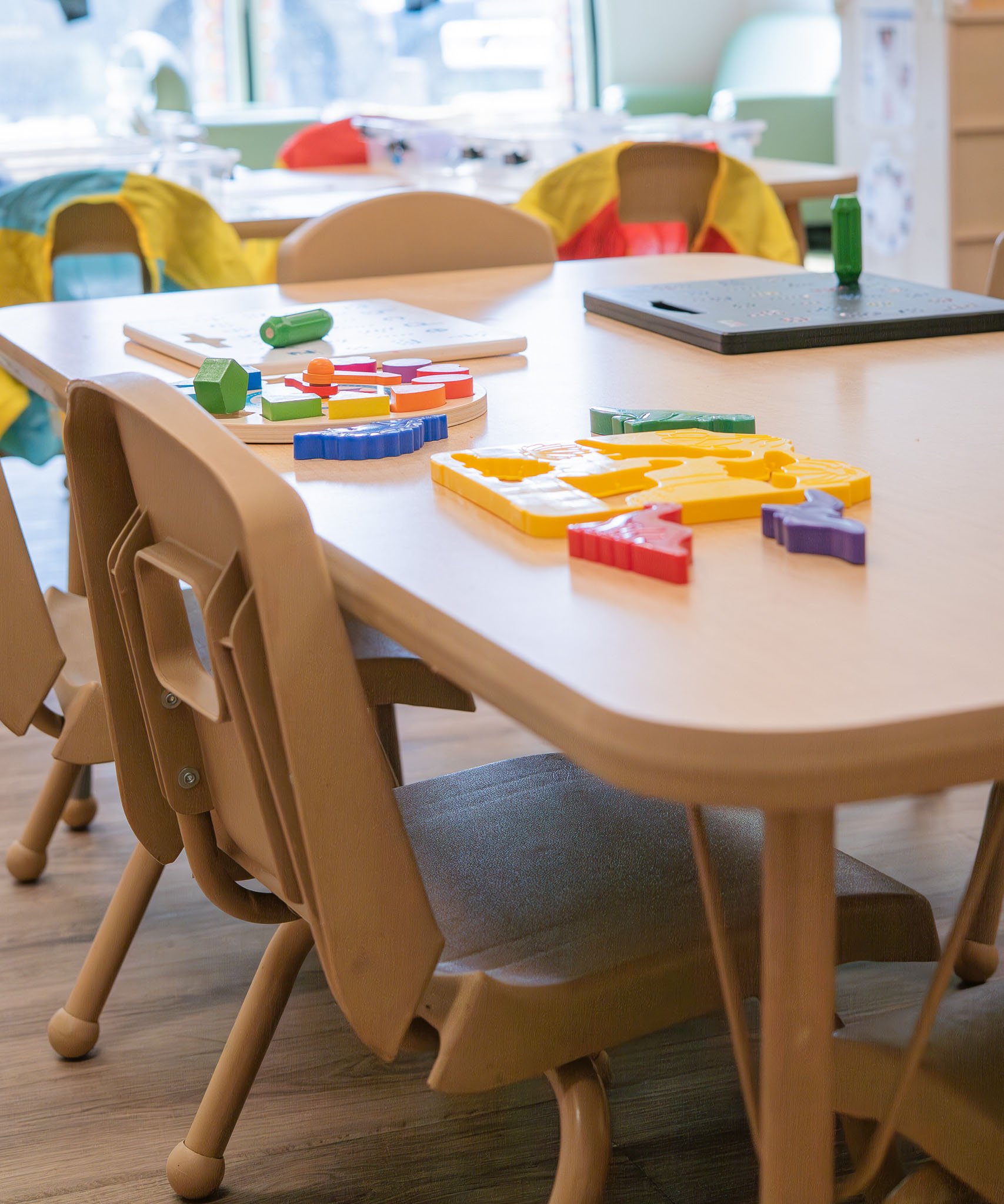 A classroom or playroom with wooden chairs and a table, several colorful wooden puzzles and toys on the table, and large windows in the background.