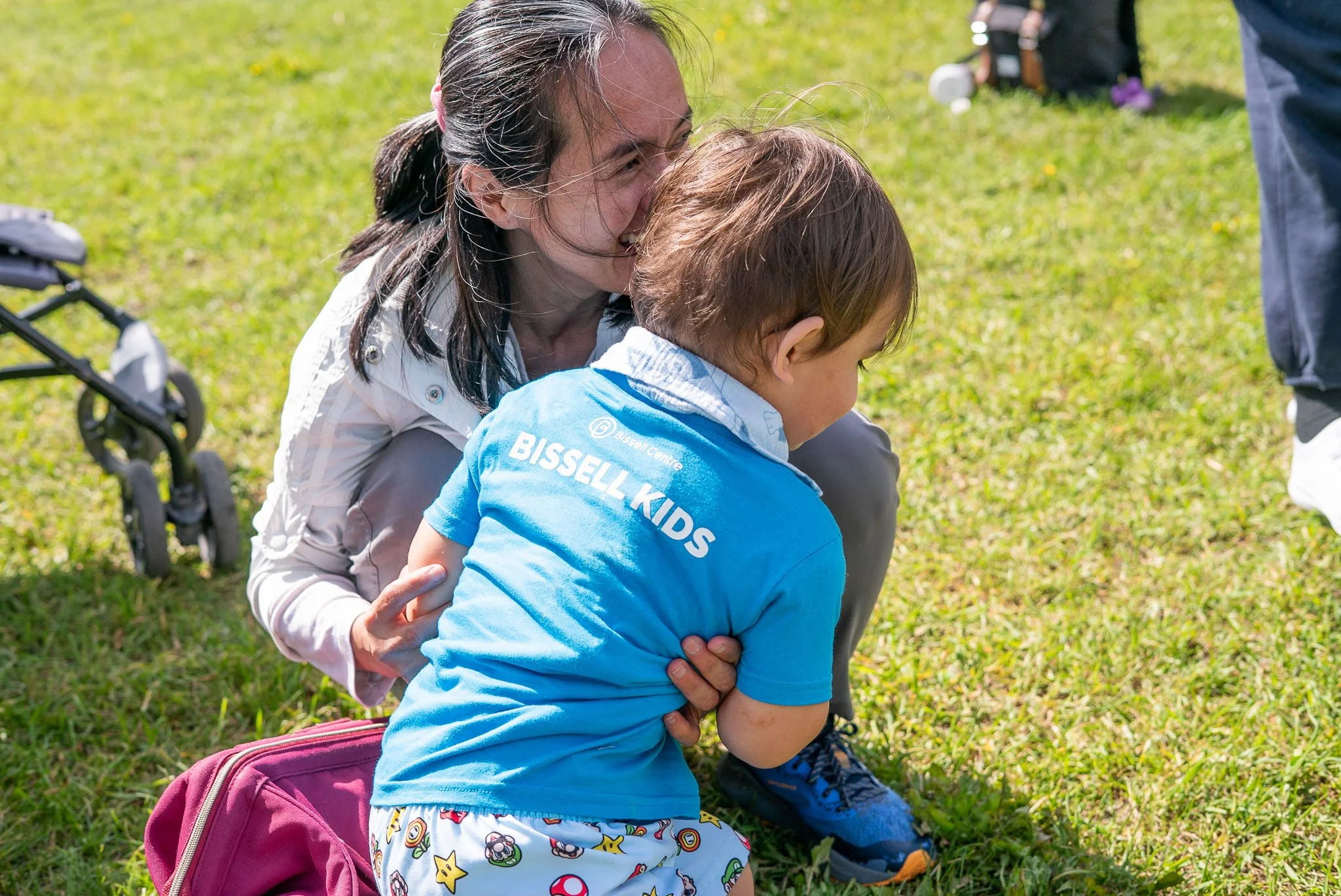 A joyful woman kneels on the grass, hugging a young boy, both smiling. They are outdoors on a sunny day, with a stroller nearby and a backpack on the ground. The woman has long dark hair and is wearing a white jacket, while the boy wears a bright blue t-shirt with cartoon and game characters on his shorts.