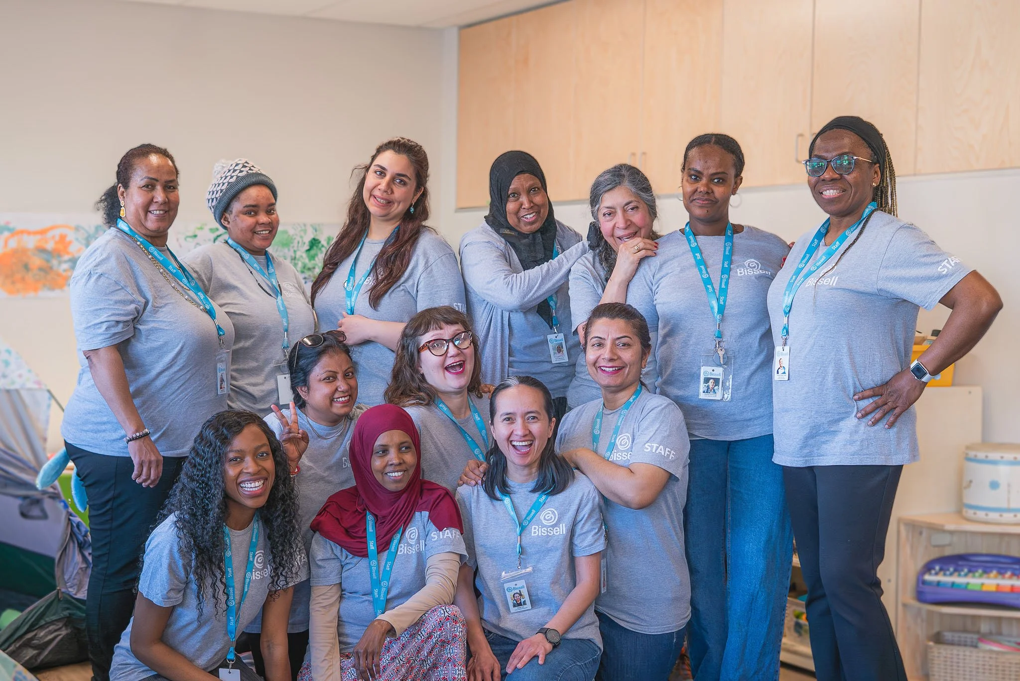 A group of diverse Early Childhood Educators smiling and posing together indoors, wearing matching blue T-shirts with the word 'Bissell Staff' written on them, all wearing blue lanyards with badges.