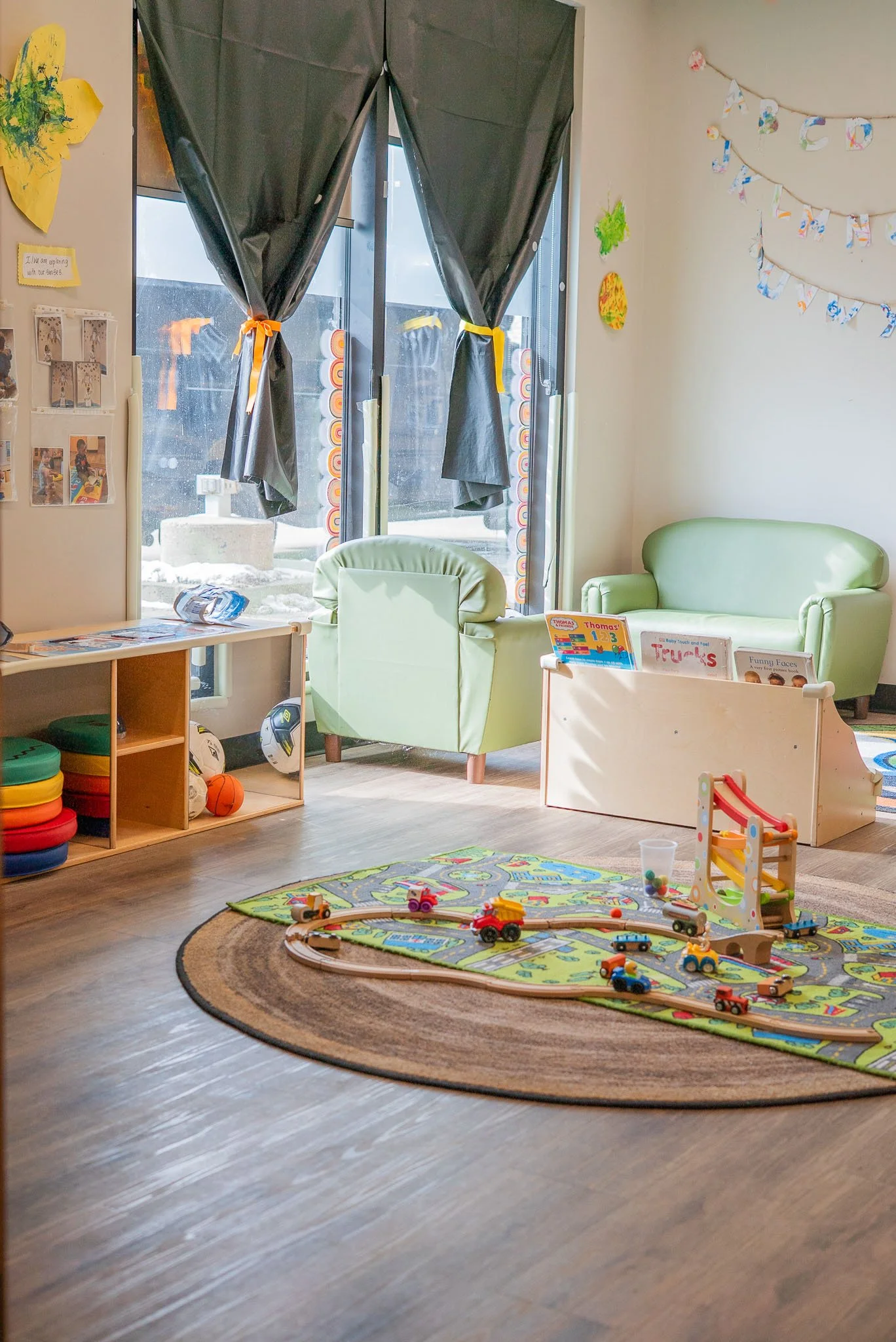 Indoor children's playroom with a train track and toy trains on a colorful rug, filled with small furniture, toys, and educational materials.