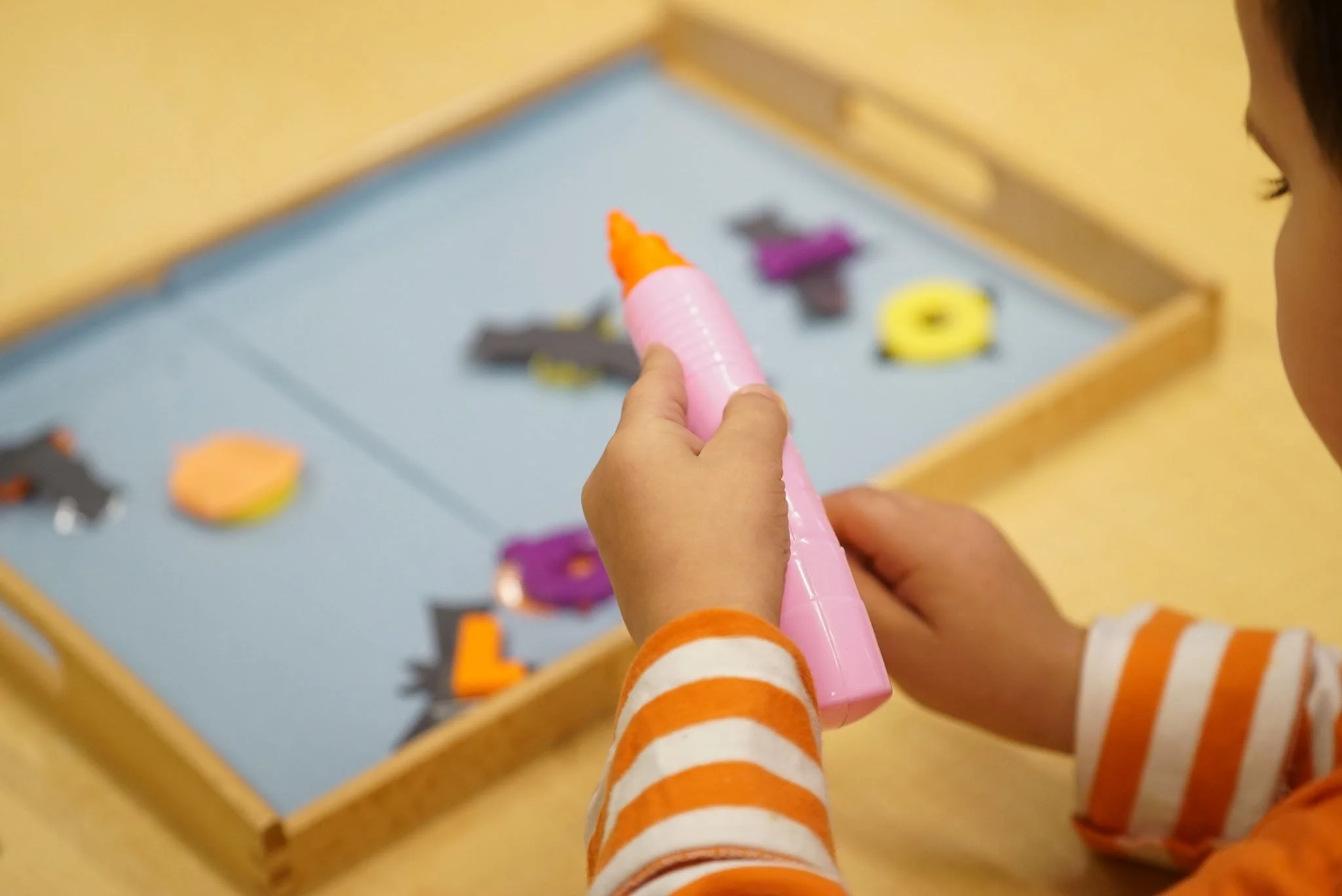 Child holding a pink marker over a wooden box filled with various colorful craft supplies.