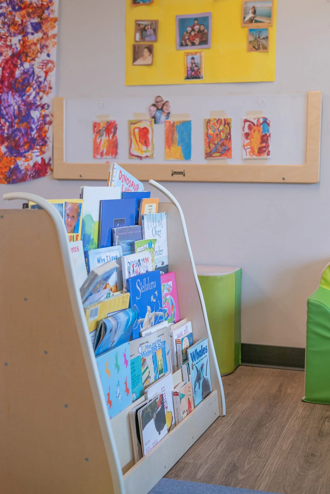 A bookshelf filled with children's books in a room with children's artwork on the walls . The wall behind has a yellow board with family photos.