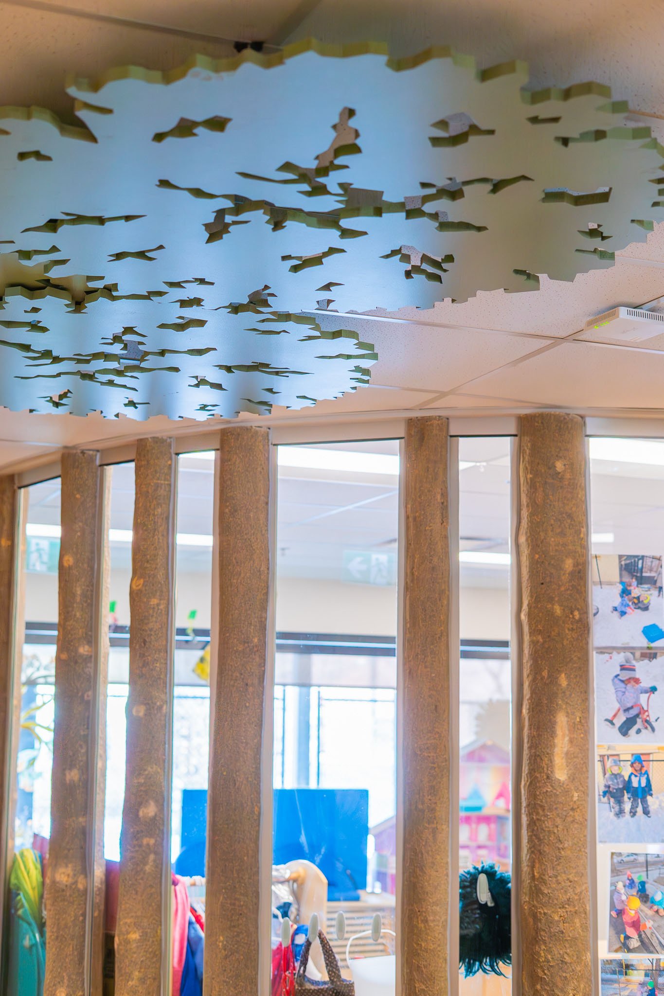 View through tree trunk style pillars of a display in a room with colorful children's artwork and toys in the background.