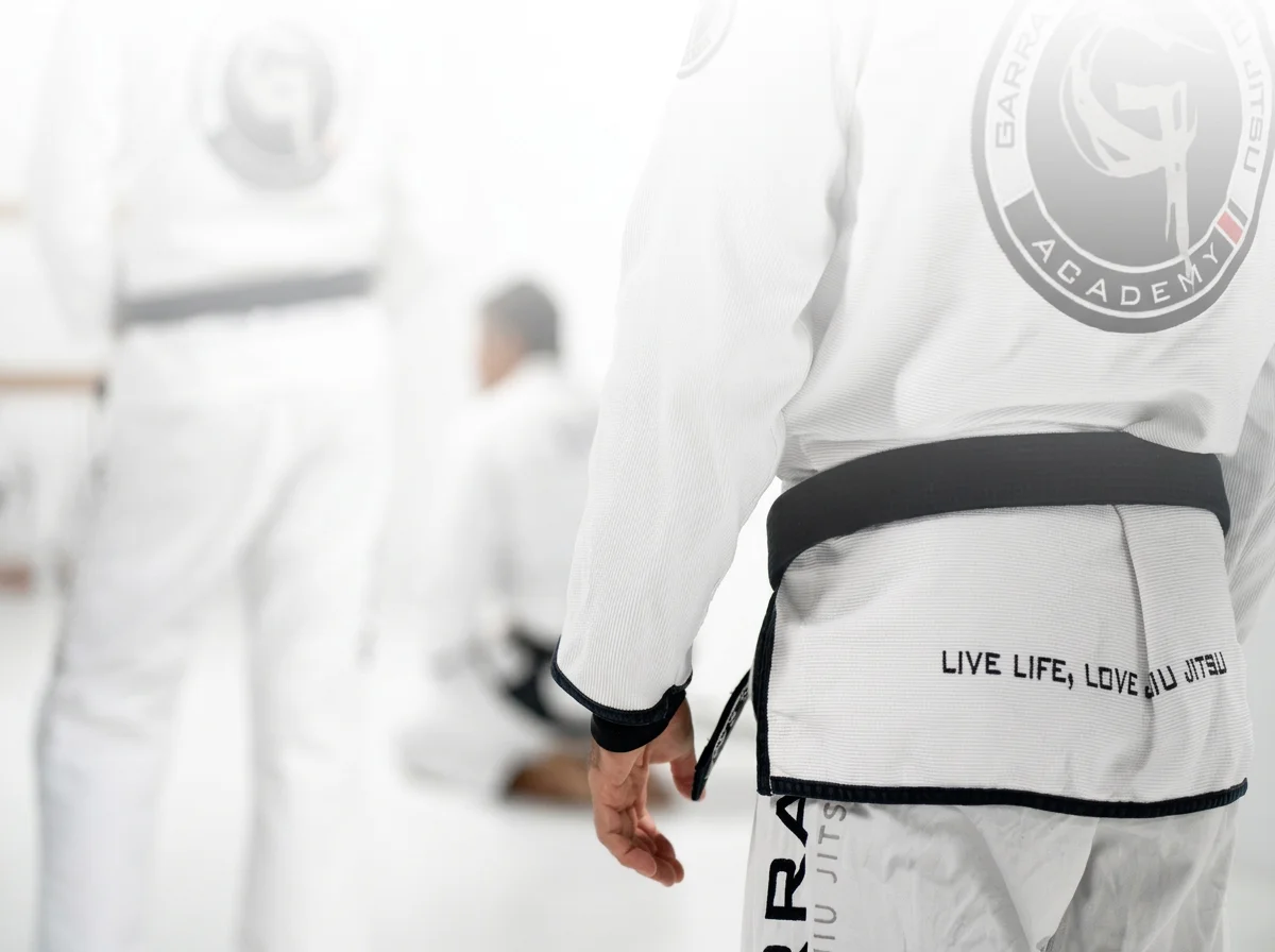 A person in a white gi with a black belt, standing in a martial arts training gym, with the gi having a patch of a martial arts academy and the words "LIVE LIFE, LOVE JIU JITSU" on the border of the belt, and other students in gis practicing in the background.