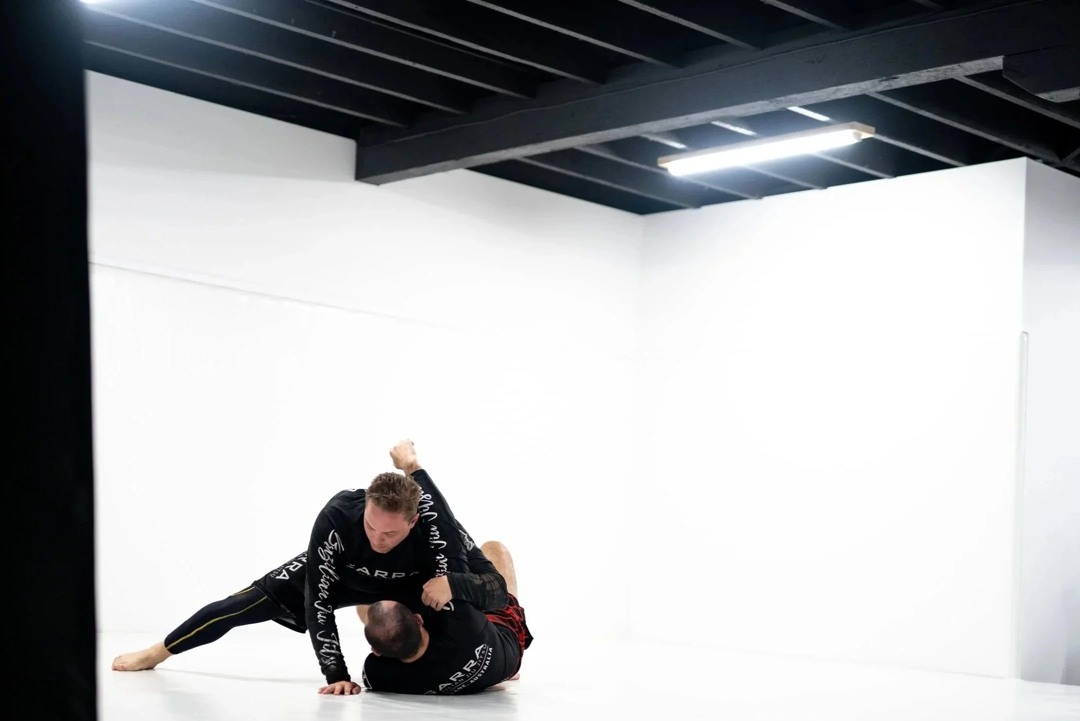 Two men practicing Brazilian Jiu-Jitsu on a white mat in a gym, one man is on top of the other who is on his back, during a grappling training session.