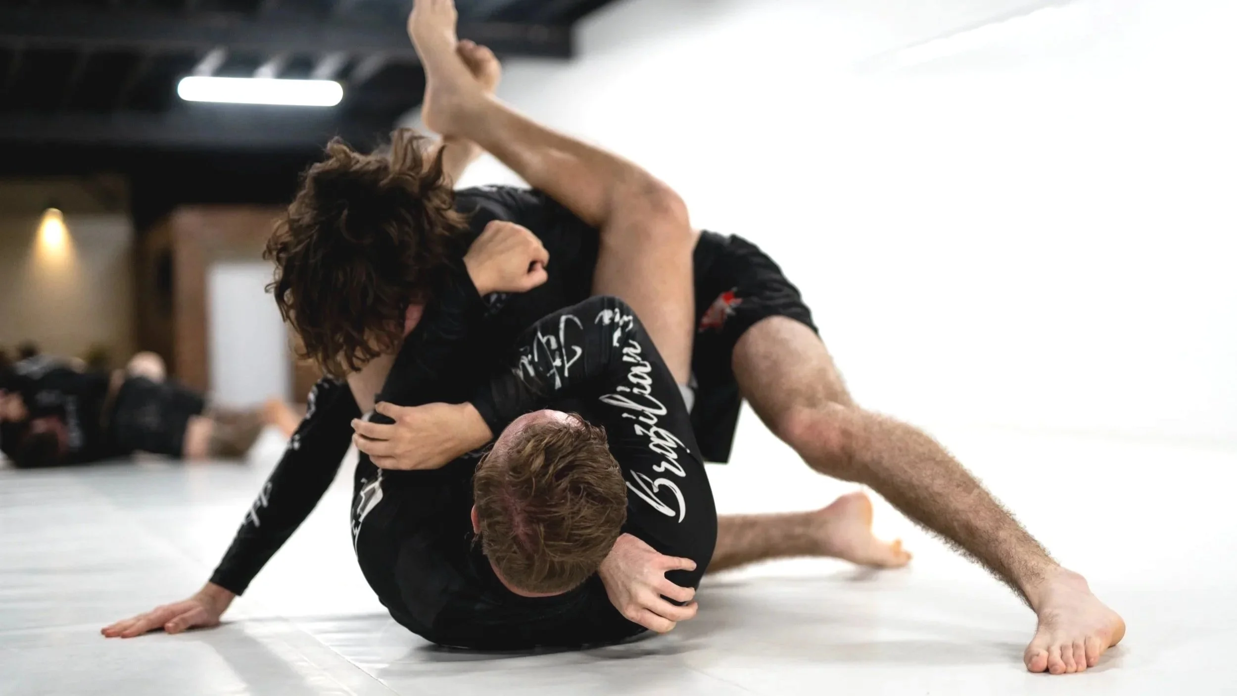 Two people practicing Brazilian Jiu-Jitsu grappling on a mat, with one person on top and the other underneath, both wearing rash guards.