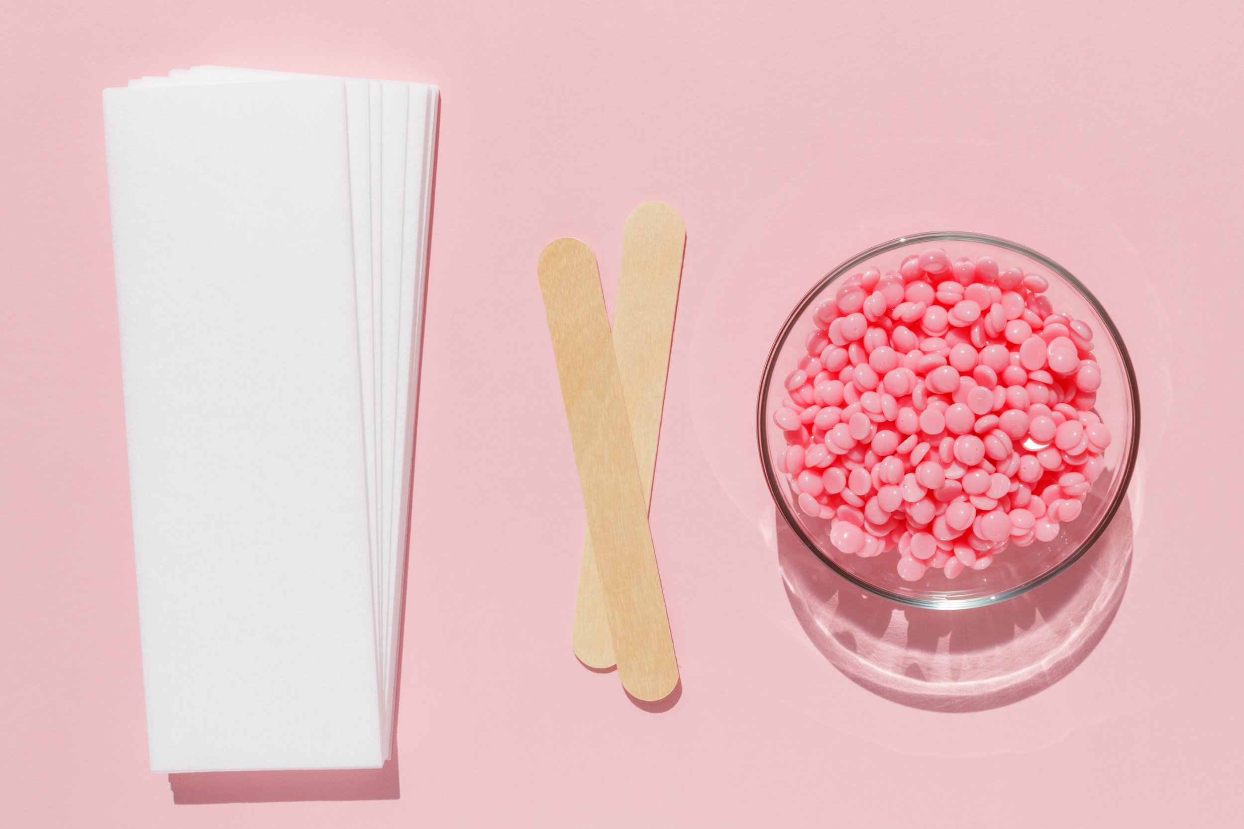 White paper straw sleeves, two wooden tongue depressors, and a glass bowl filled with pink wax beads on a pink background.