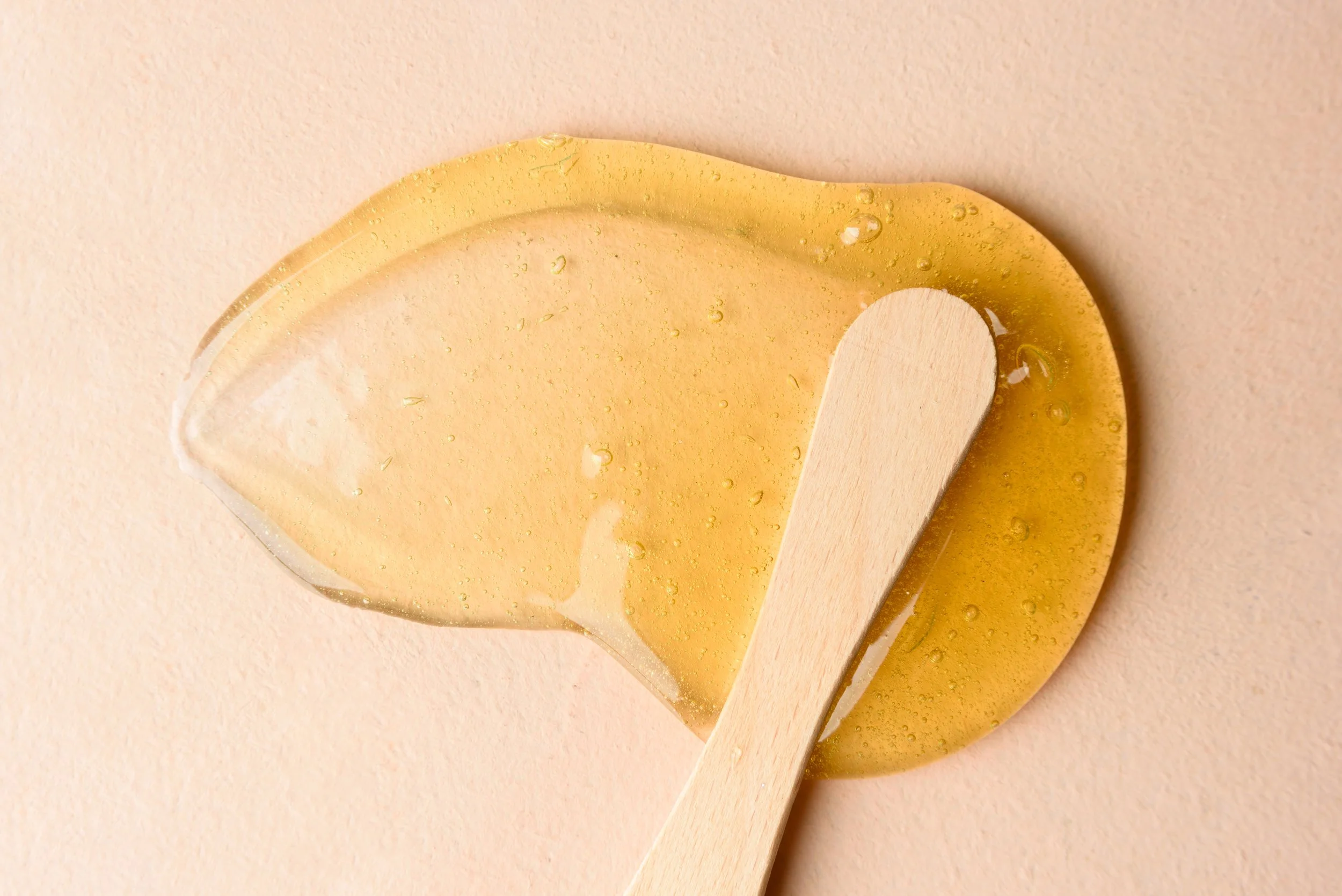 Close-up of a clear yellow wax on a small spatula, with tiny air bubbles, against a soft beige background.