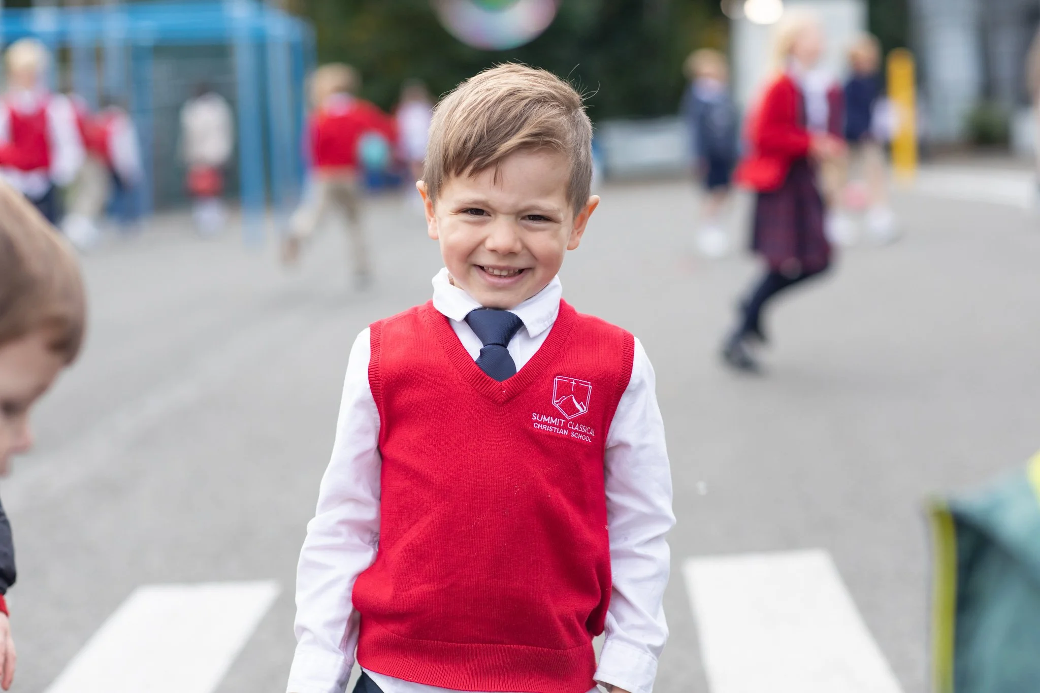 Young Summit student in a red sweater vest and tie smiling on the playground