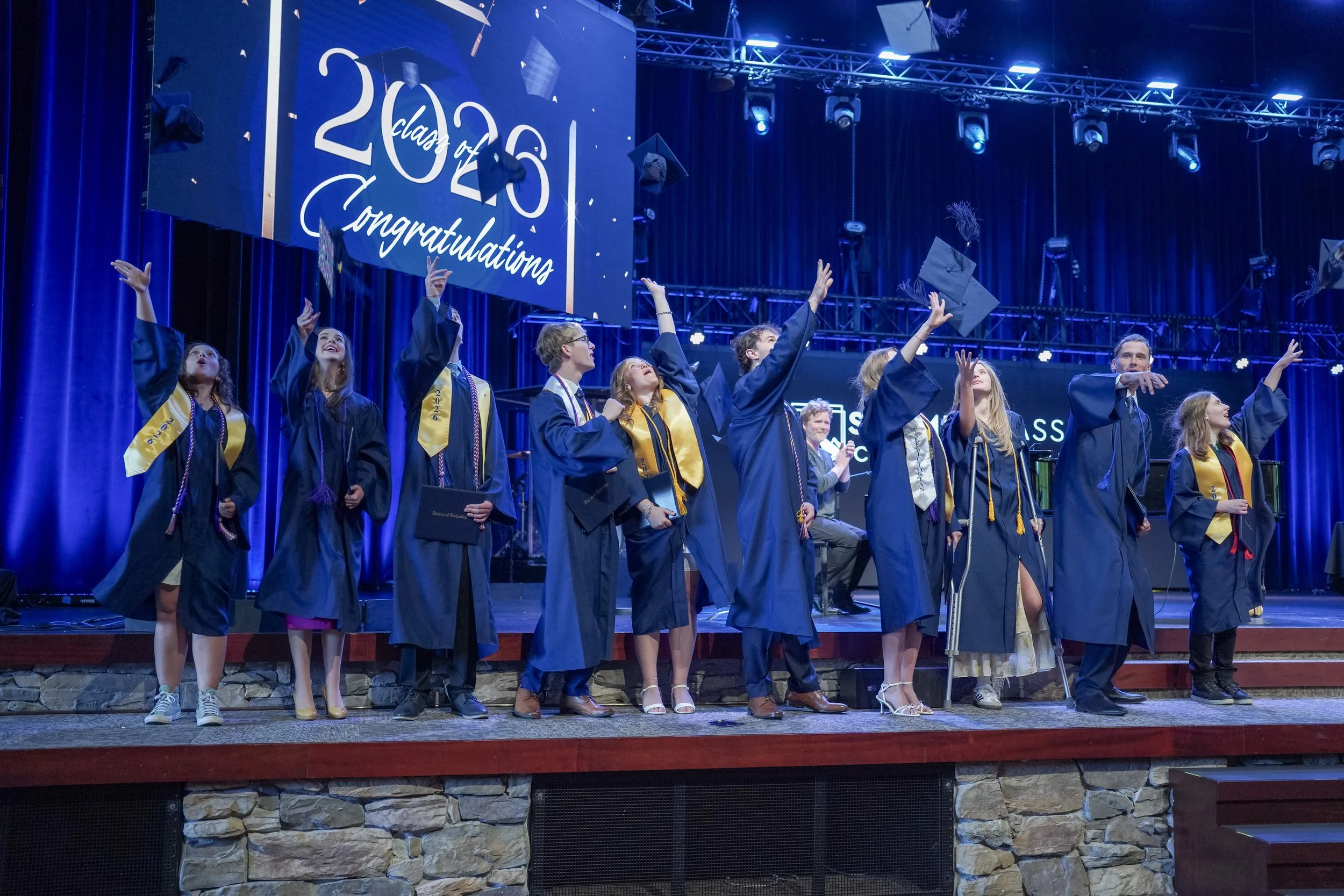 Graduates in blue caps and gowns celebrate onstage beneath a 2023 congratulations banner.