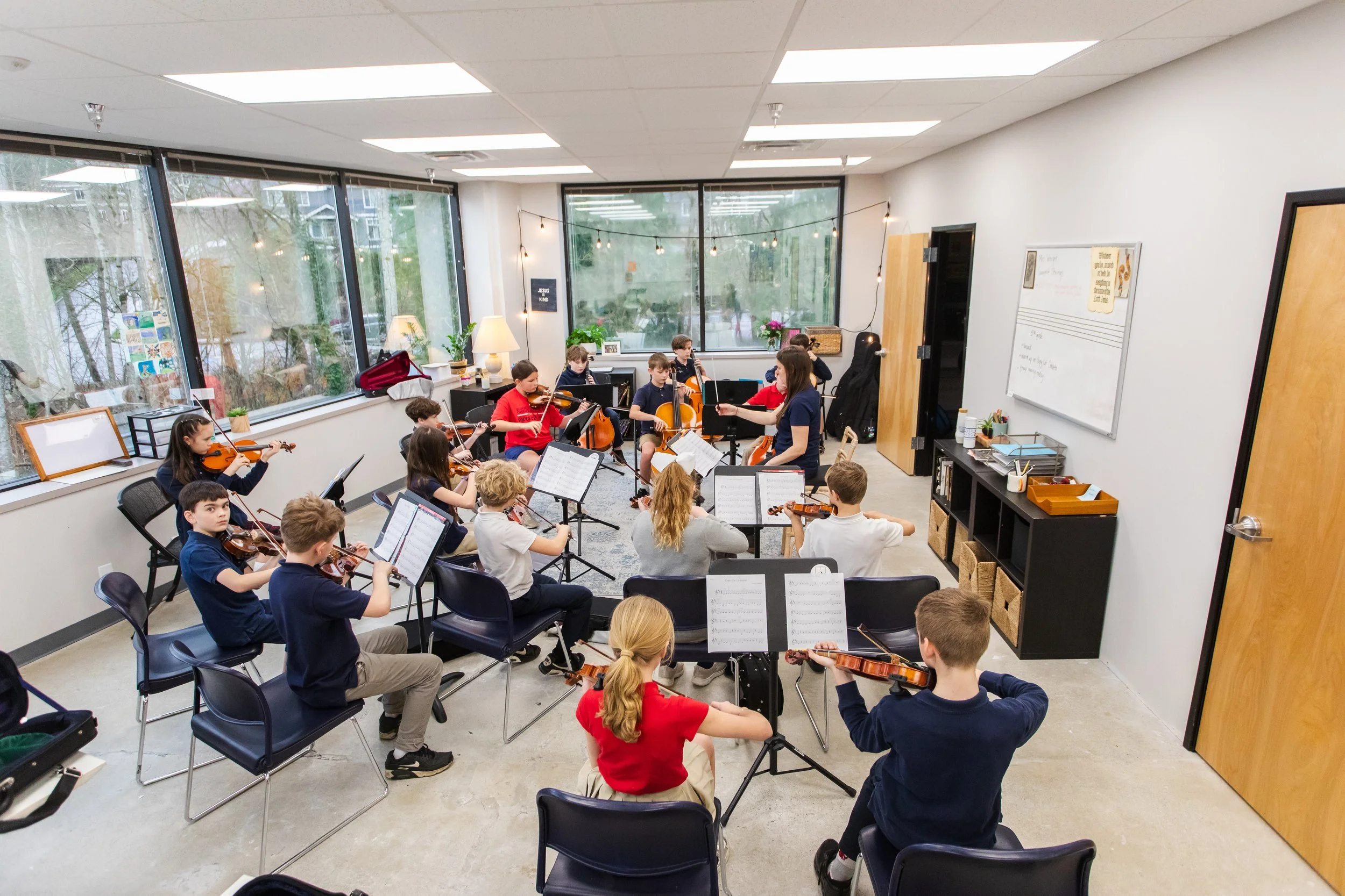 Children practicing string instruments with a teacher in a bright classroom orchestra rehearsal.
