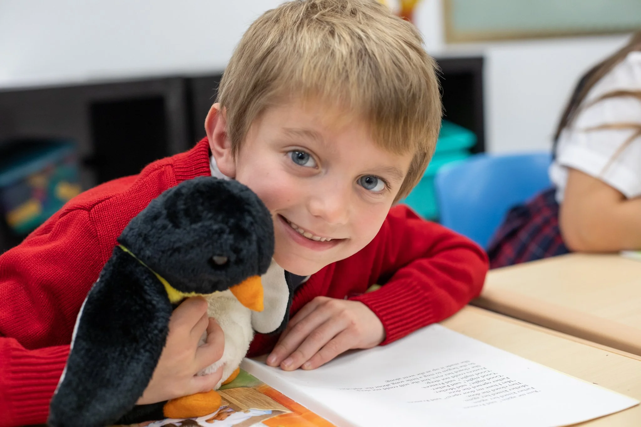 Lower School student in a red sweater smiling with a stuffed penguin at his desk