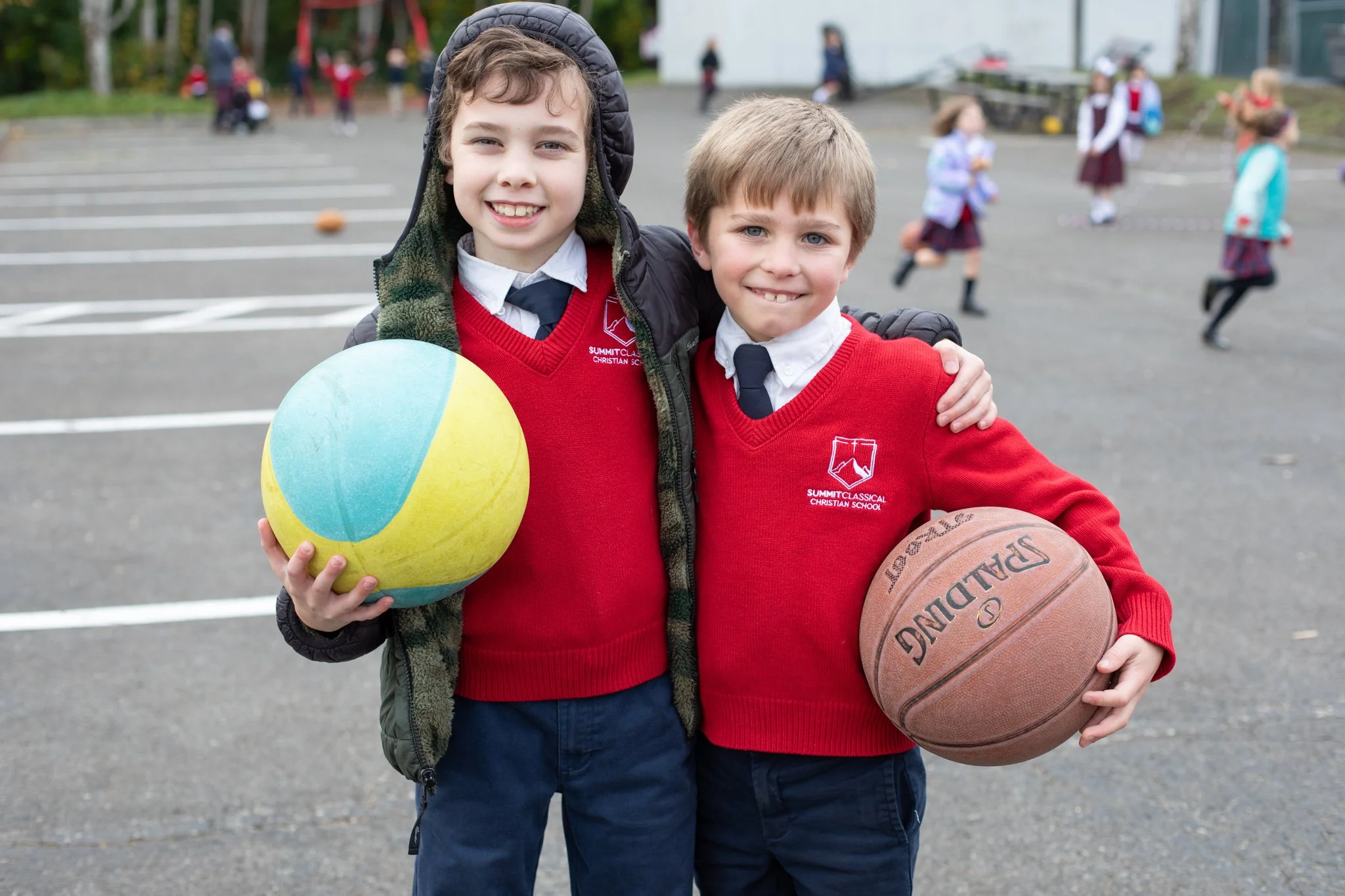 Two smiling schoolboys in red uniforms holding a colorful ball and basketball on a school playground.
