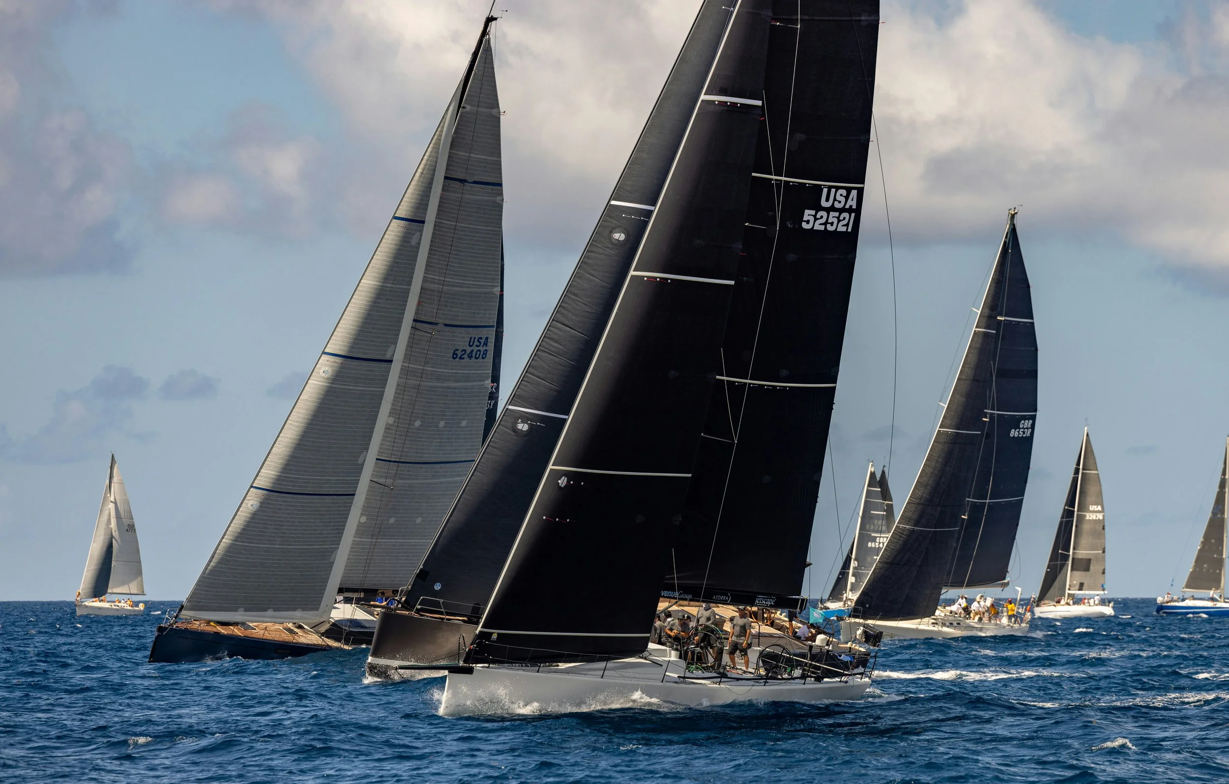 Multiple sailboats racing on the water with mostly black sails and some with gray and white sails, under a partly cloudy sky.