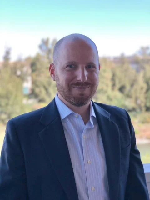 A man with a shaved head and beard wearing a dark blue suit jacket and a light blue dress shirt, standing outdoors with blurred trees in the background.