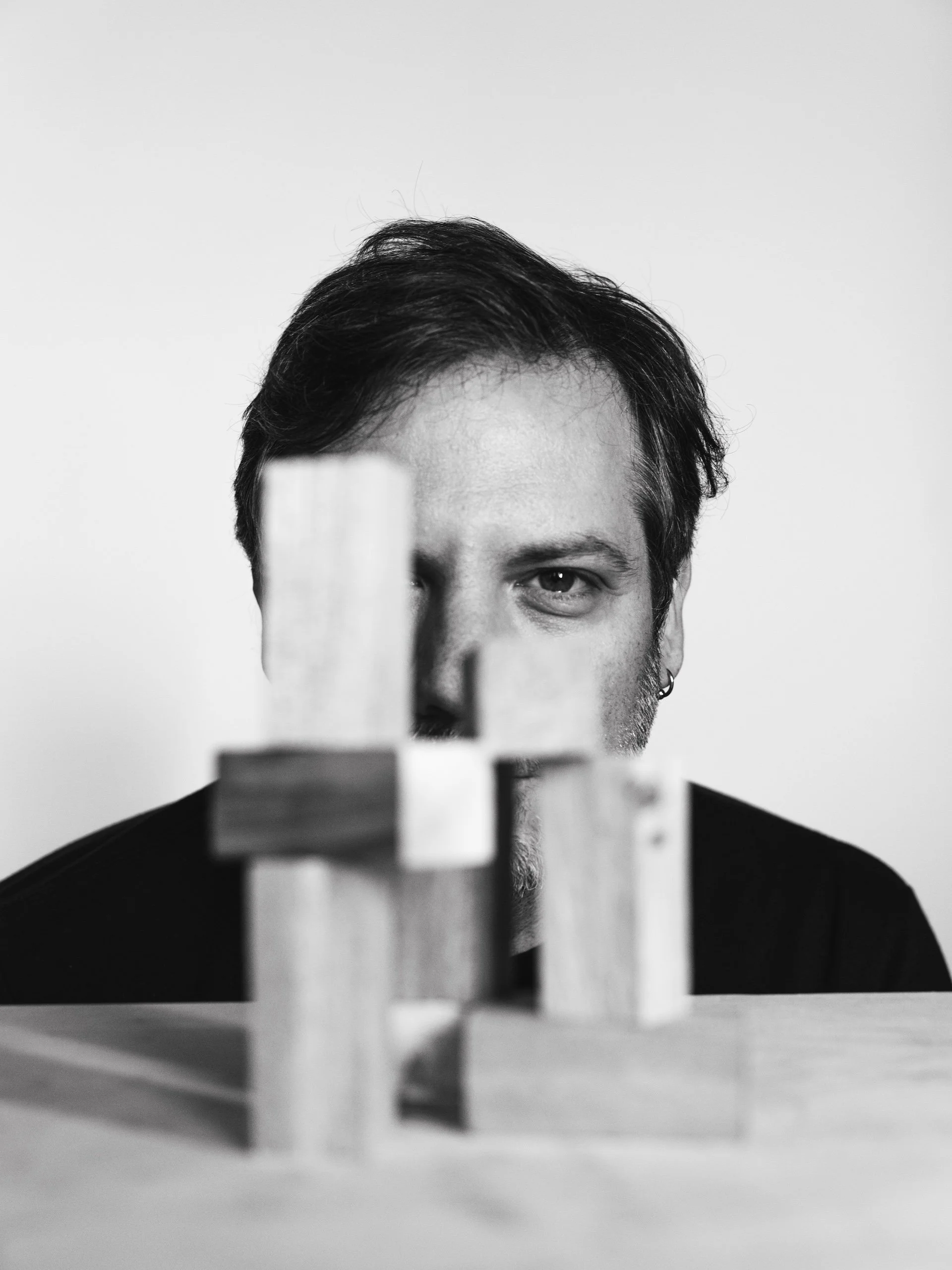 Persona con cabello oscuro, solo la mitad superior de la cara visible, en un fondo gris claro, frente a bloques de madera apilados en primer plano.
