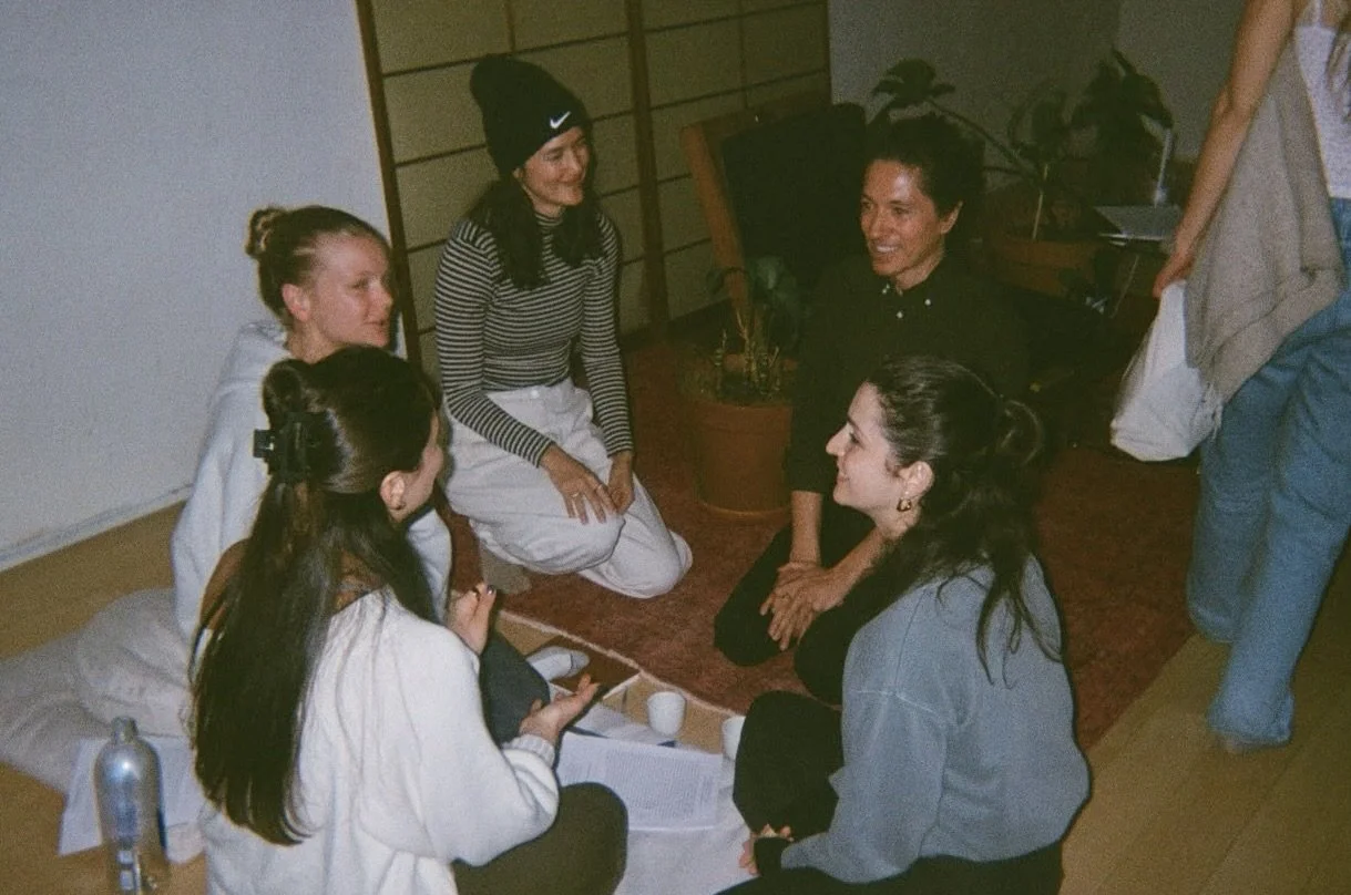 Grupo de seis personas conversando en un ambiente interior, algunas están sentadas en el suelo y otras están de rodillas, en el centro hay una mujer de cabello oscuro y ropa negra, y otra mujer con cabello recogido y vestido gris.