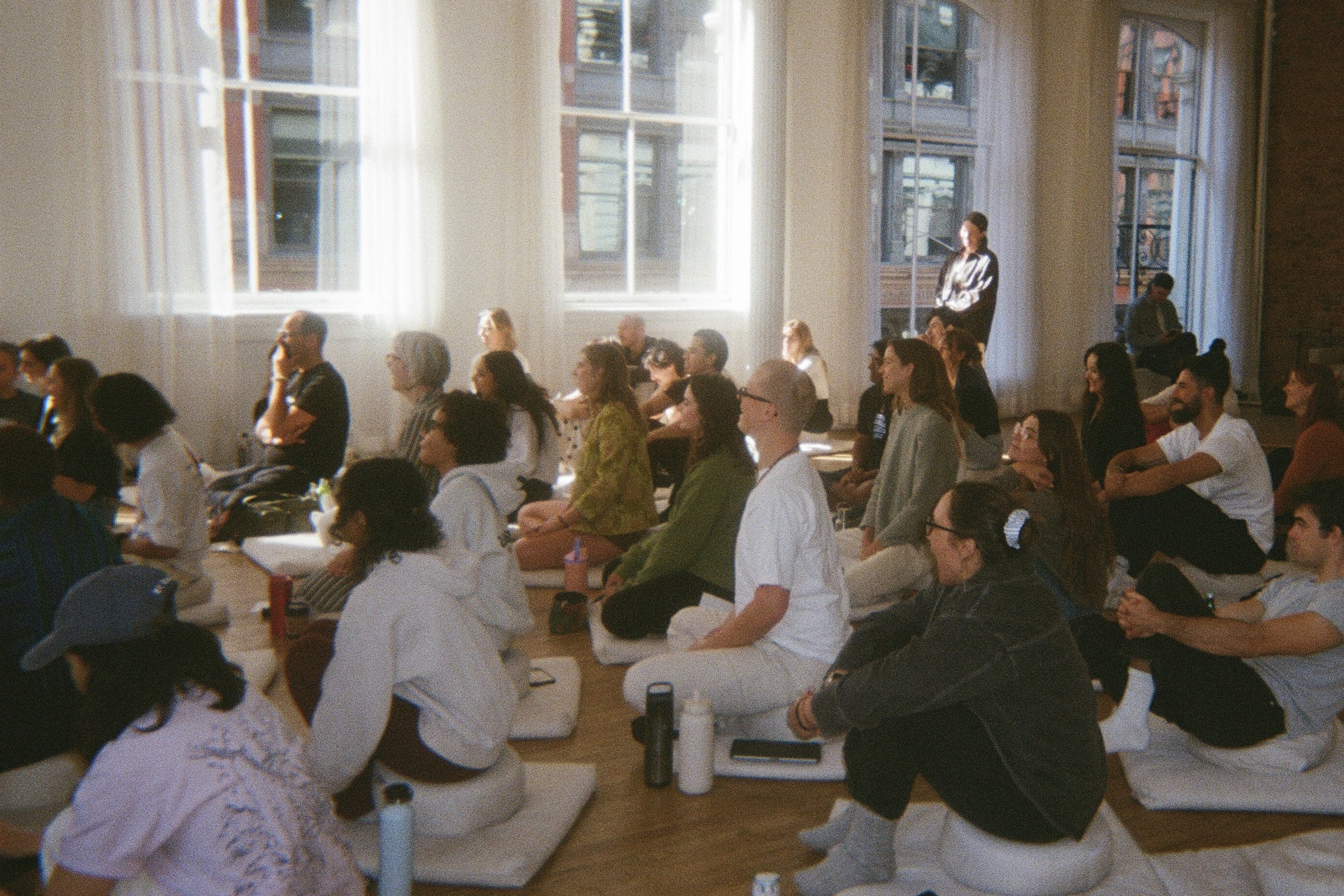 Personas sentadas en la sala durante la formacion de profesores de meditacion, algunas personas tomando notas y otras escuchando, ambiente luminoso y tranquilo.