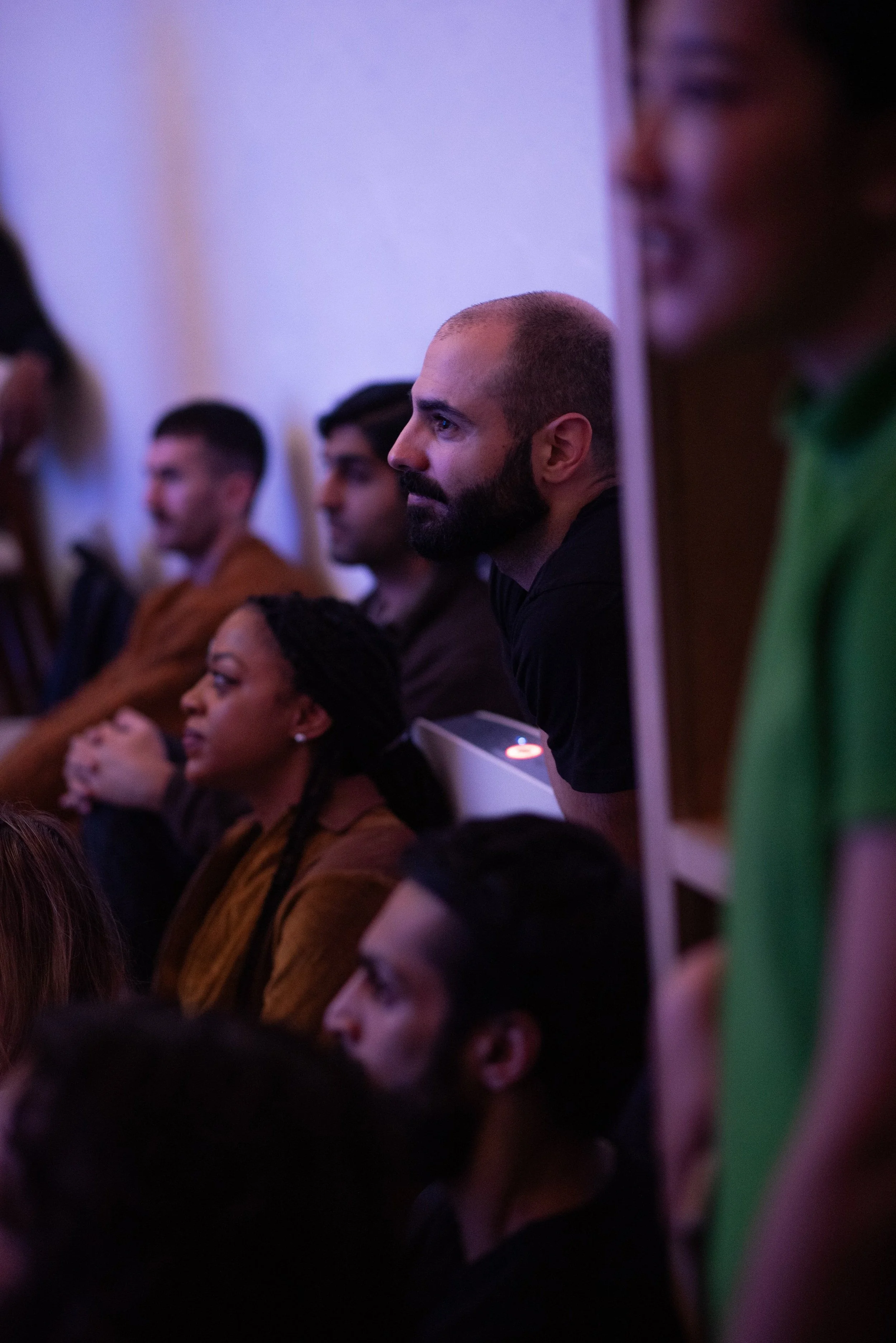 Grupo de personas sentadas en una reunión o conferencia, con enfoque en un hombre calvo de barba, vistiendo una camiseta negra, observando atentamente.