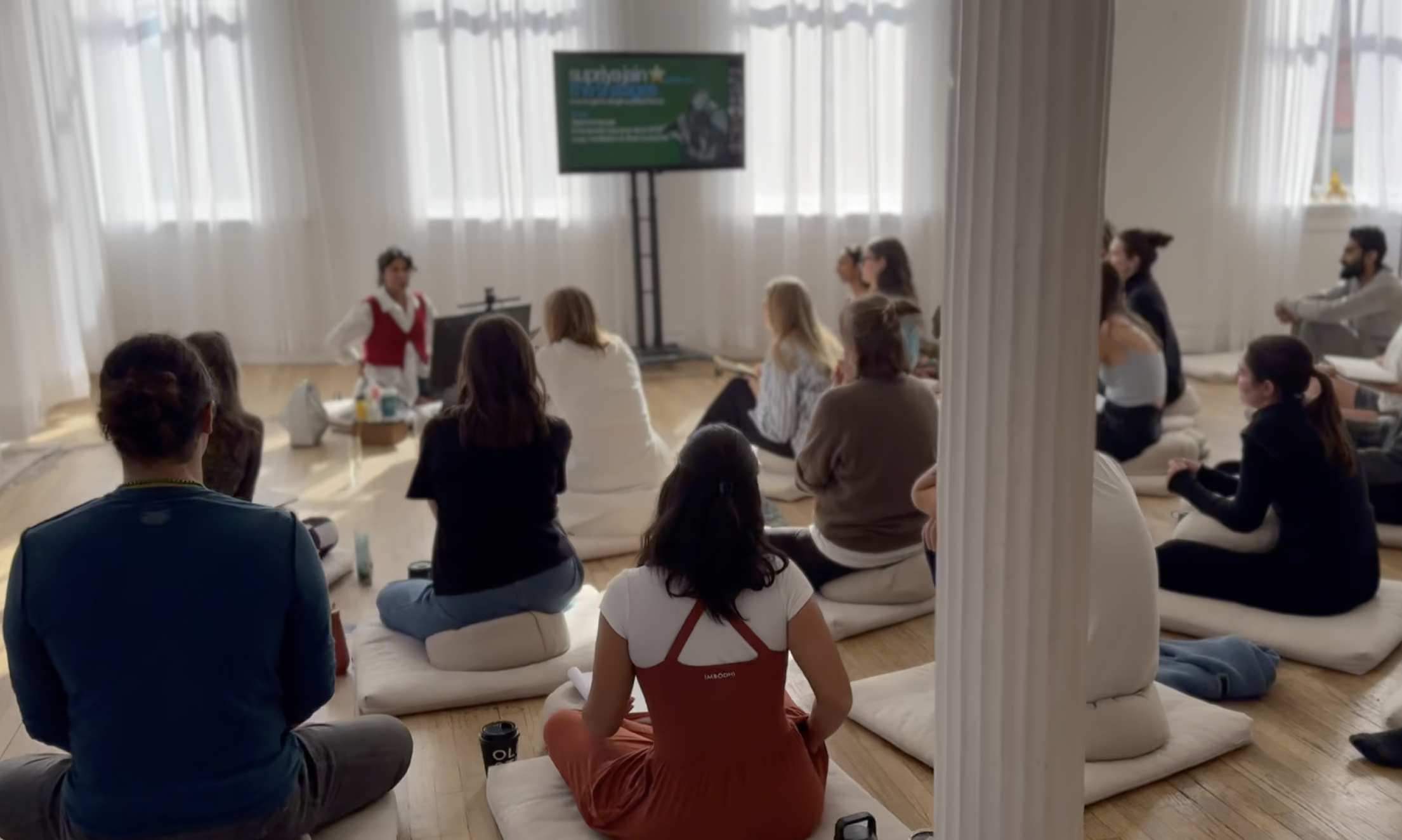 Grupo de personas en una sala, participando en una sesión de meditación, sentados en cojines en el suelo, frente a una instructora.