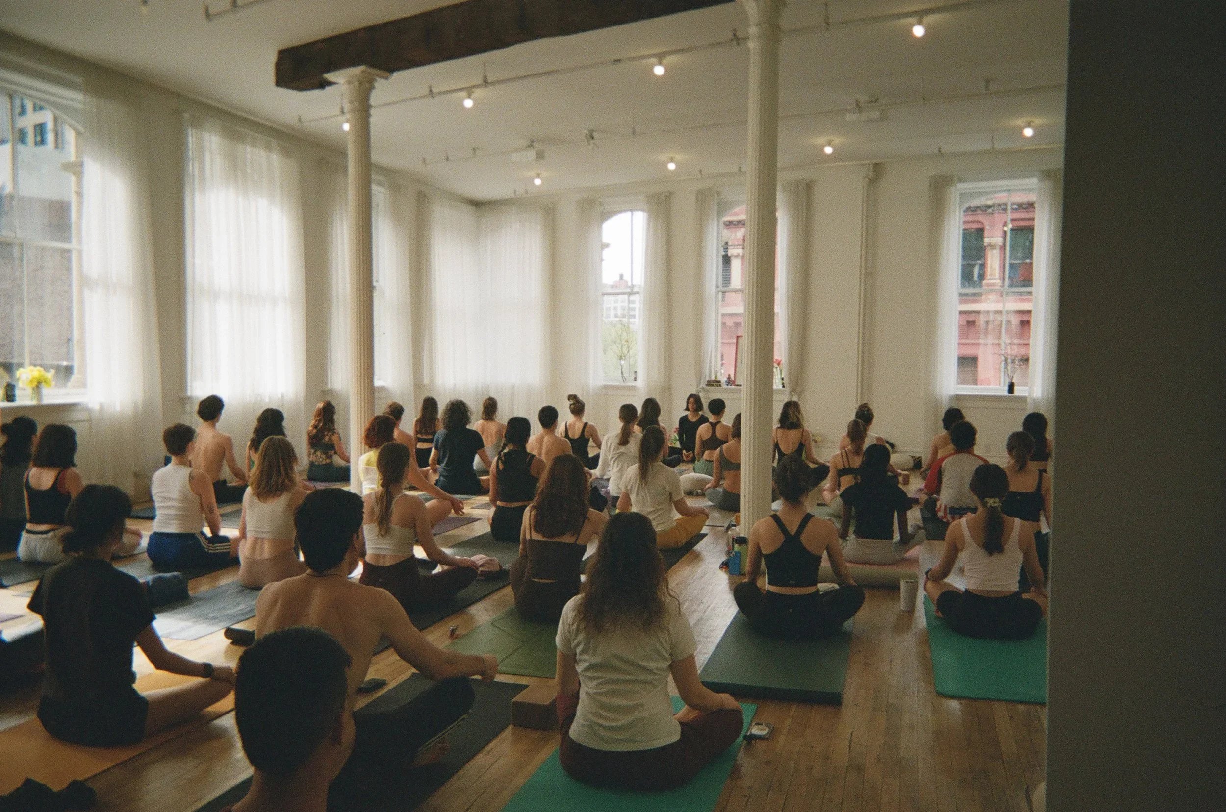 Grupo de personas participando en una clase de yoga en un espacio grande con ventanas grandes y cortinas blancas, y decoraciones sencillas.
